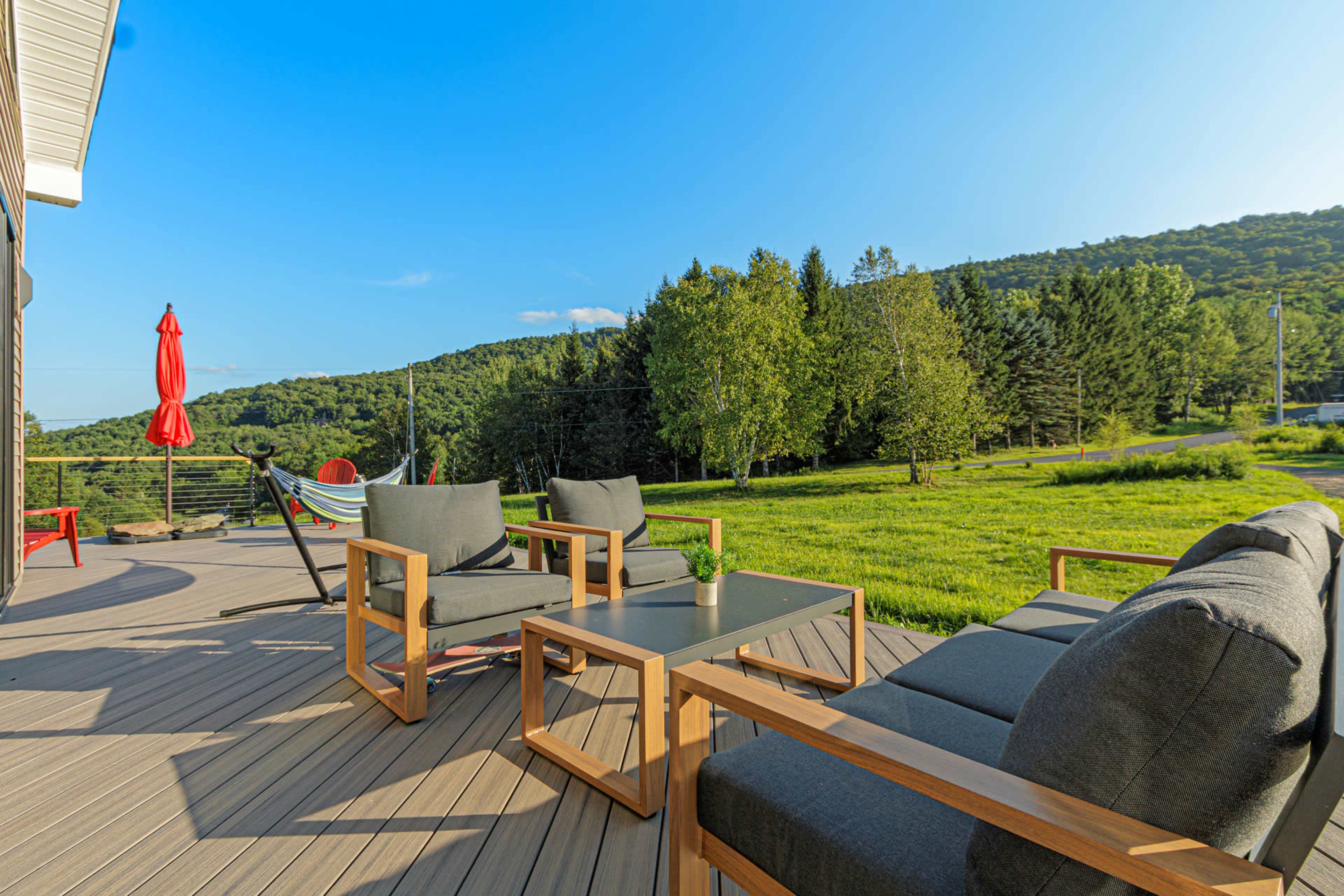 A patio area featuring wooden furniture, a hammock, and a red umbrella, overlooking a grassy field and wooded hills.