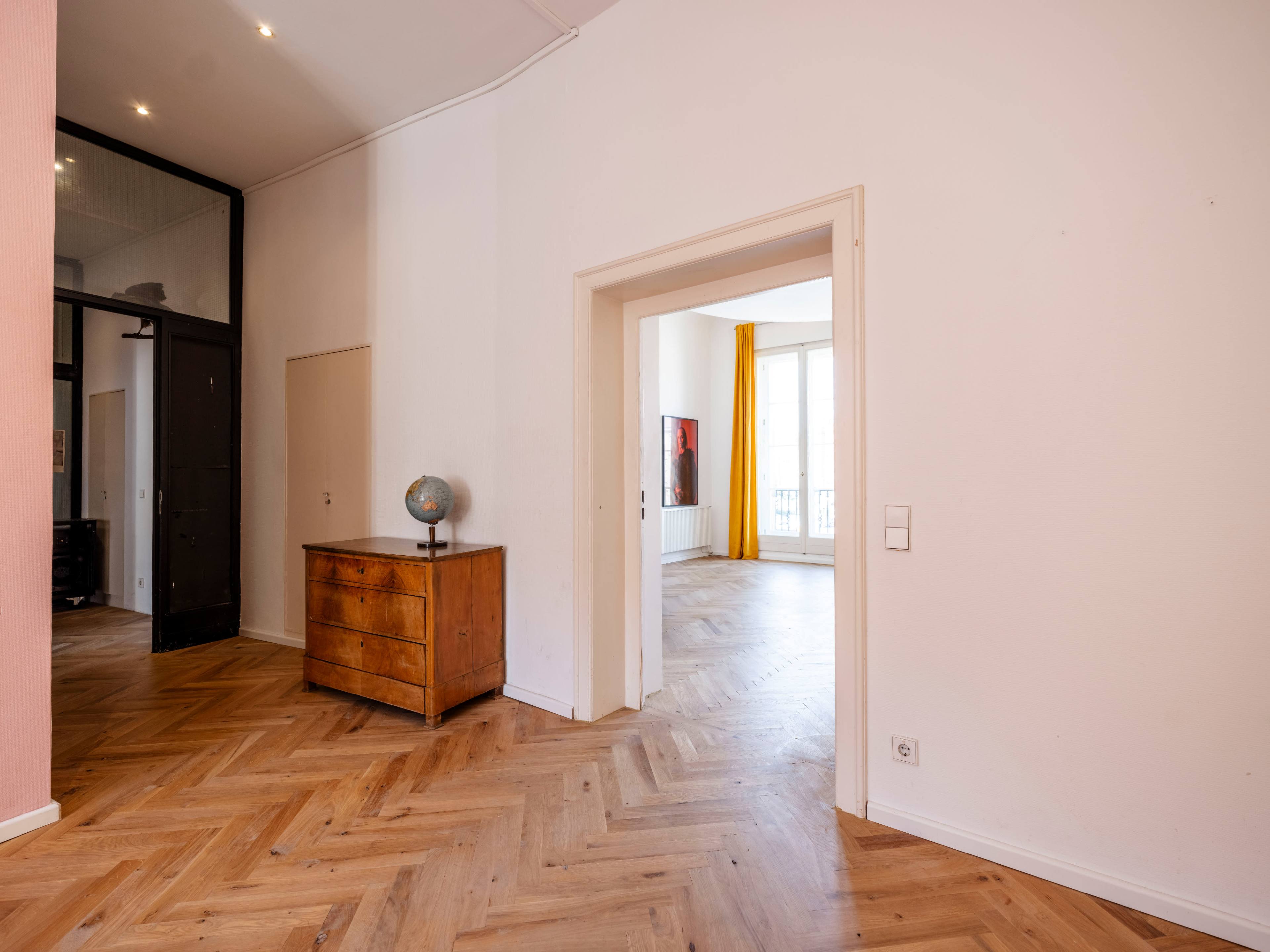 The image shows a spacious entrance area with a wooden dresser, an open doorway leading to a bright room with large windows, and herringbone-patterned wooden flooring.