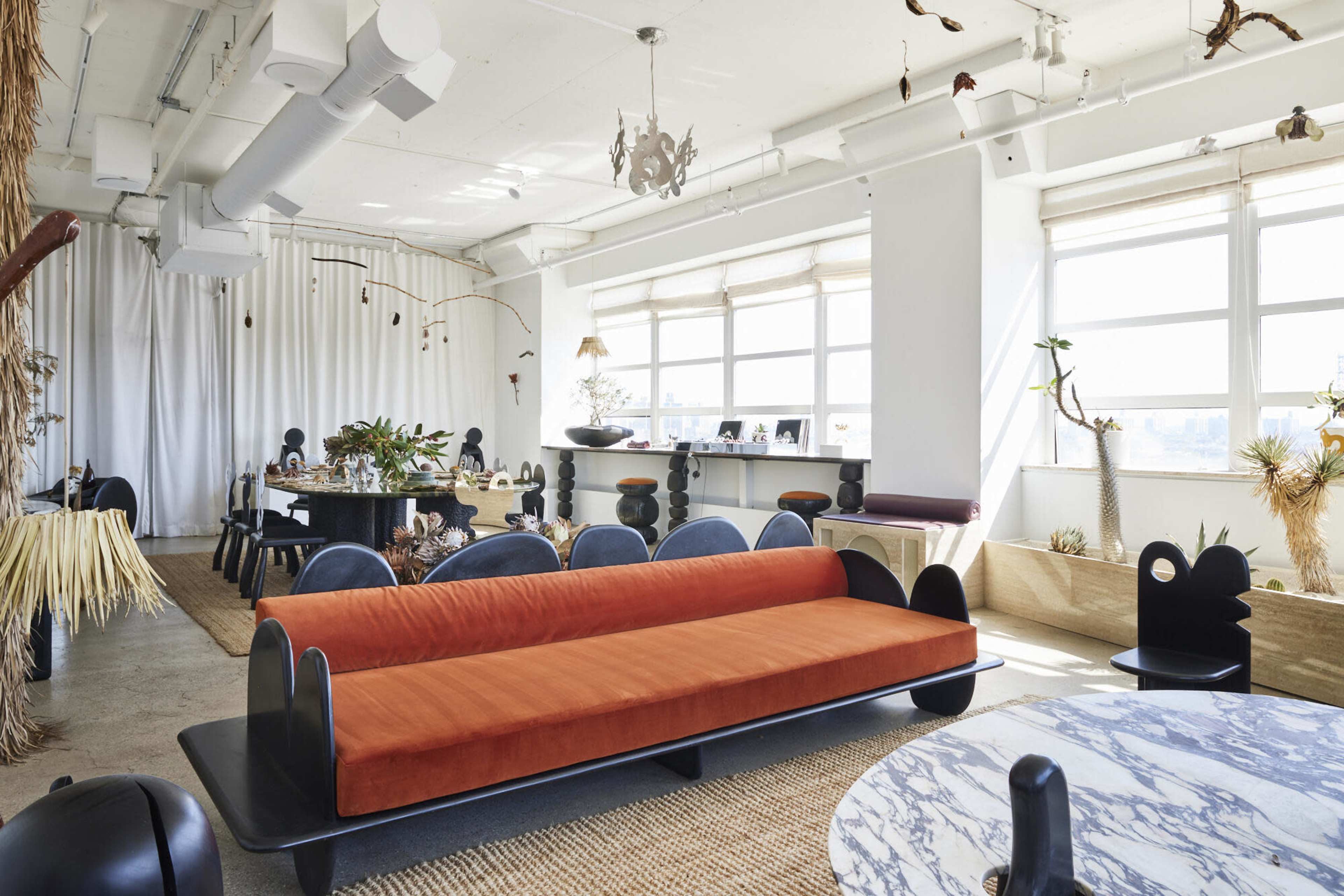 The image shows a spacious modern interior featuring a black and orange sofa, a dining table with decorative items, and large windows allowing natural light.