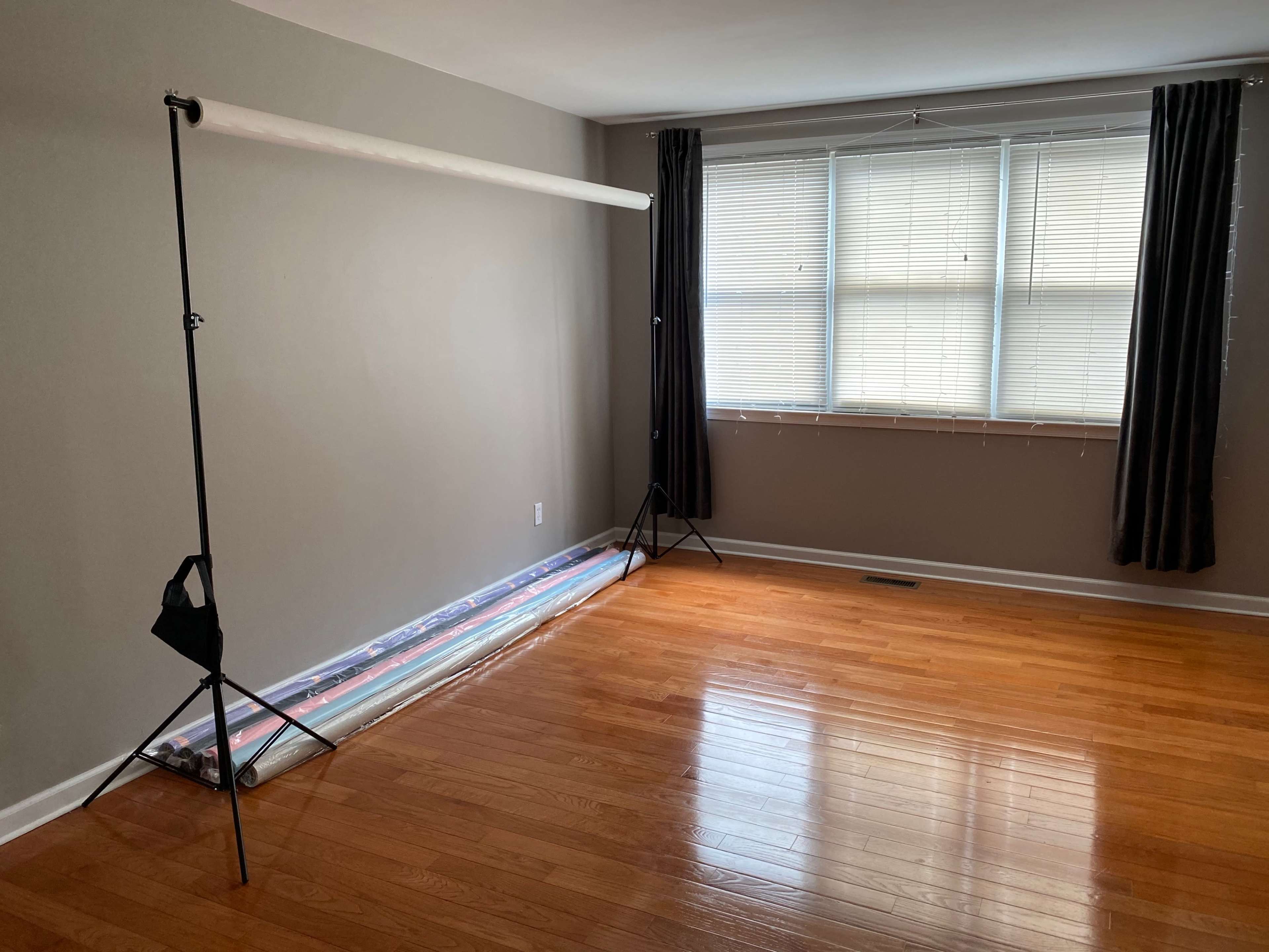 An empty room with polished wooden flooring, a backdrop stand with white fabric, and a roll of backdrop material on the floor near a large window with horizontal blinds.