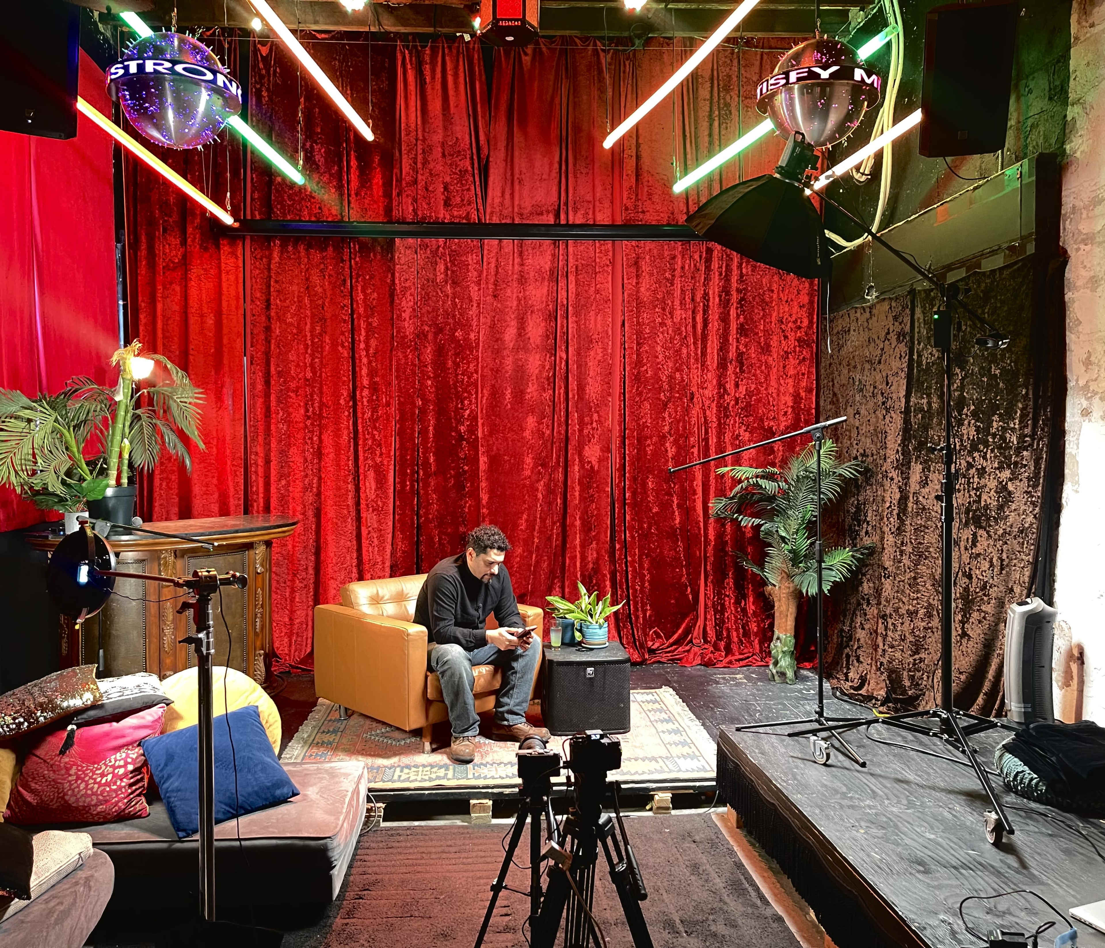 A man sits on a brown couch in a vibrant studio setting with red curtains, colorful lights, and filming equipment.