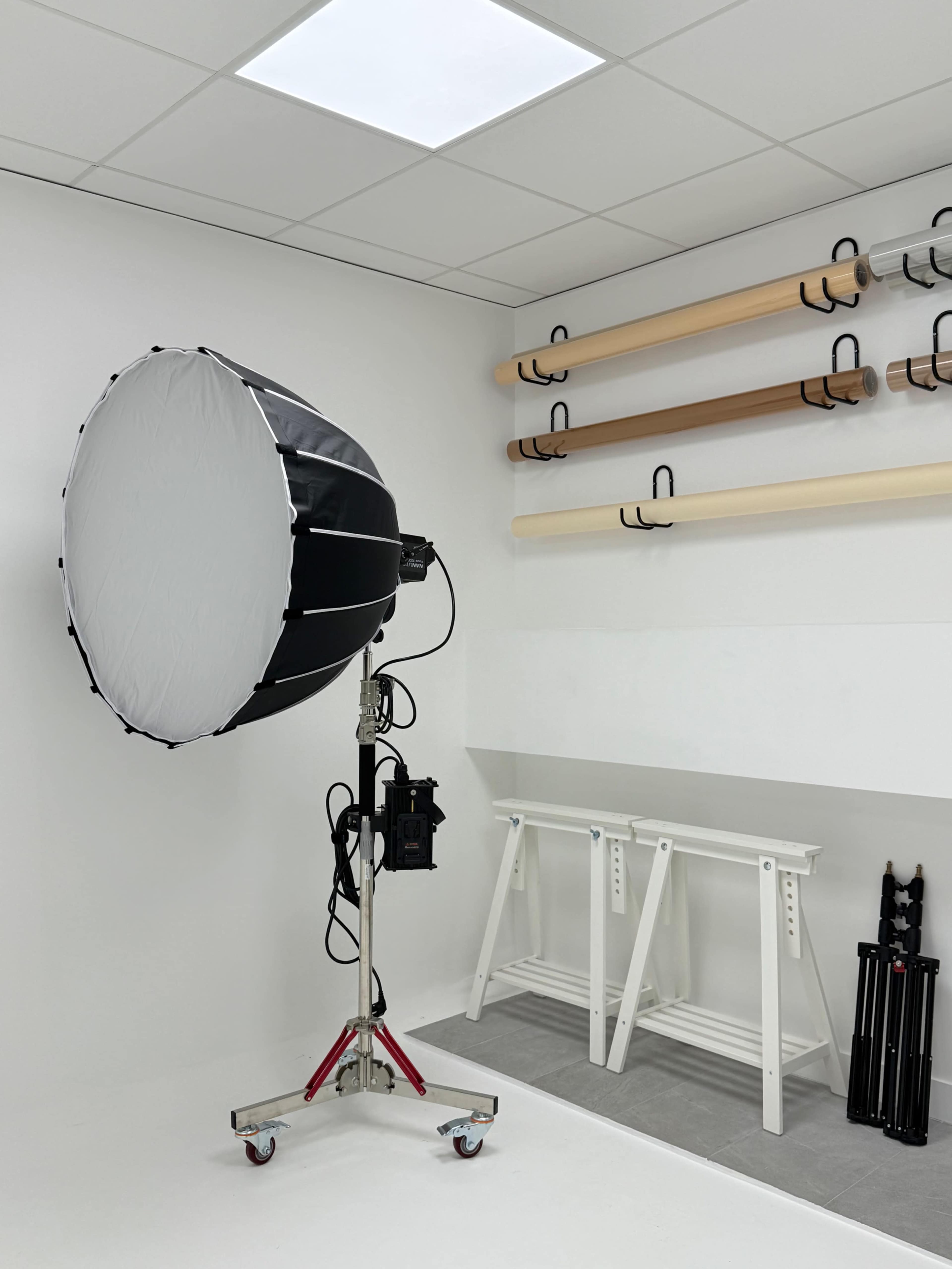 A large photography softbox on a stand is positioned in a well-lit, minimalistic studio with wooden shelves and a set of sawhorses.