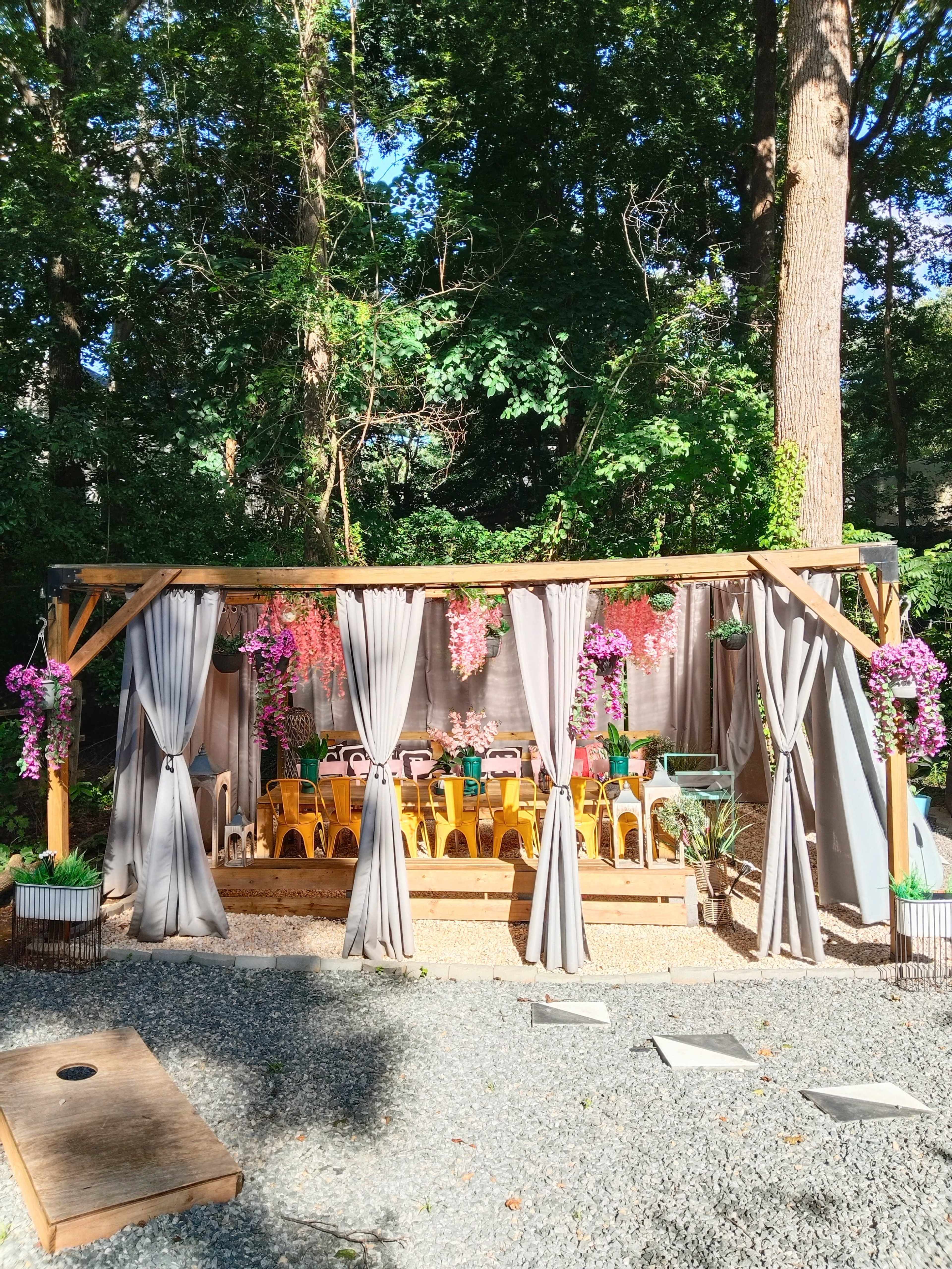 The image shows a outdoor gathering space with a wooden pergola adorned with colorful flowers and grey curtains, surrounded by gravel and trees.