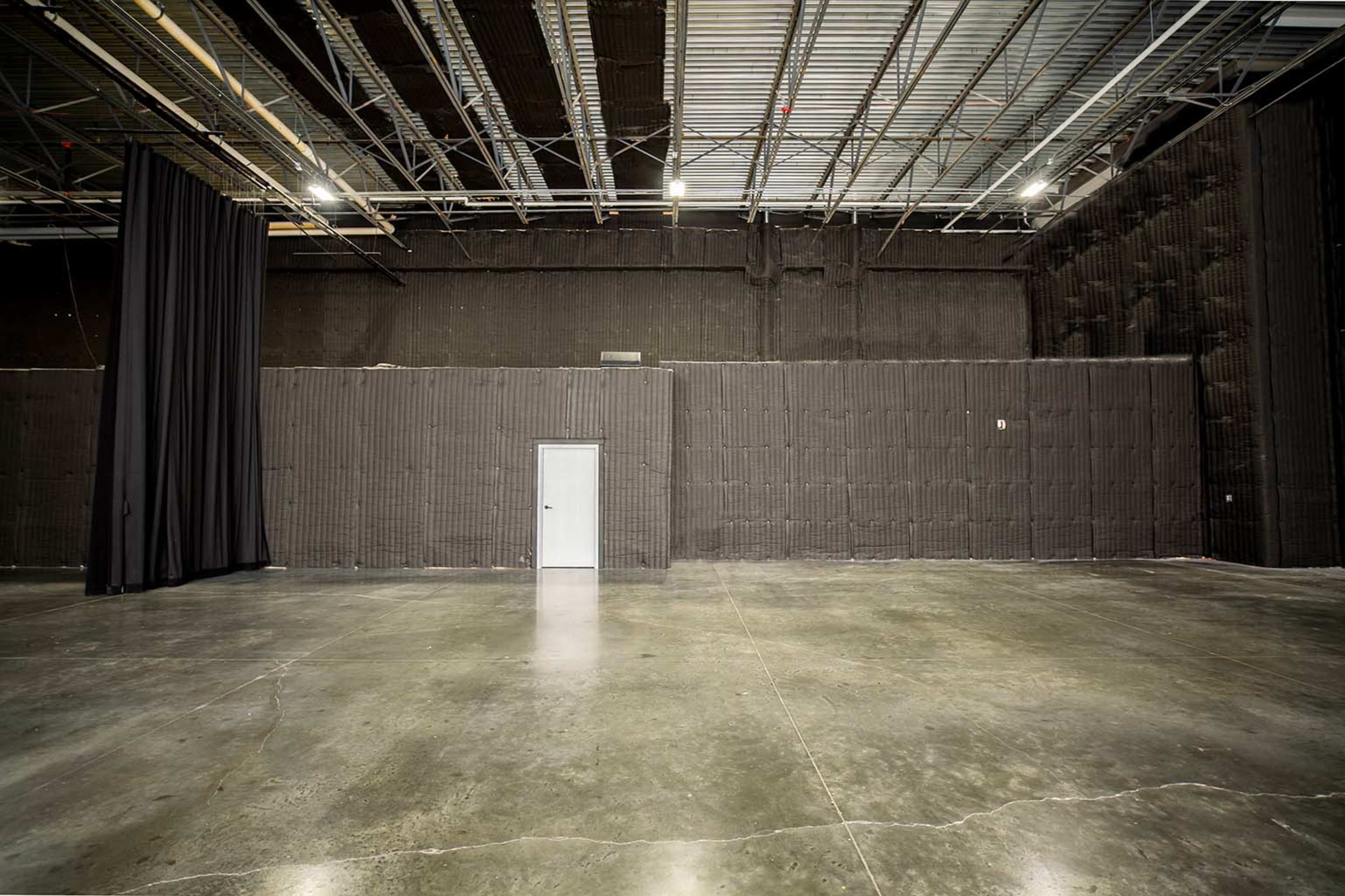A large, empty warehouse space with a concrete floor and a single door against a black soundproofed wall.
