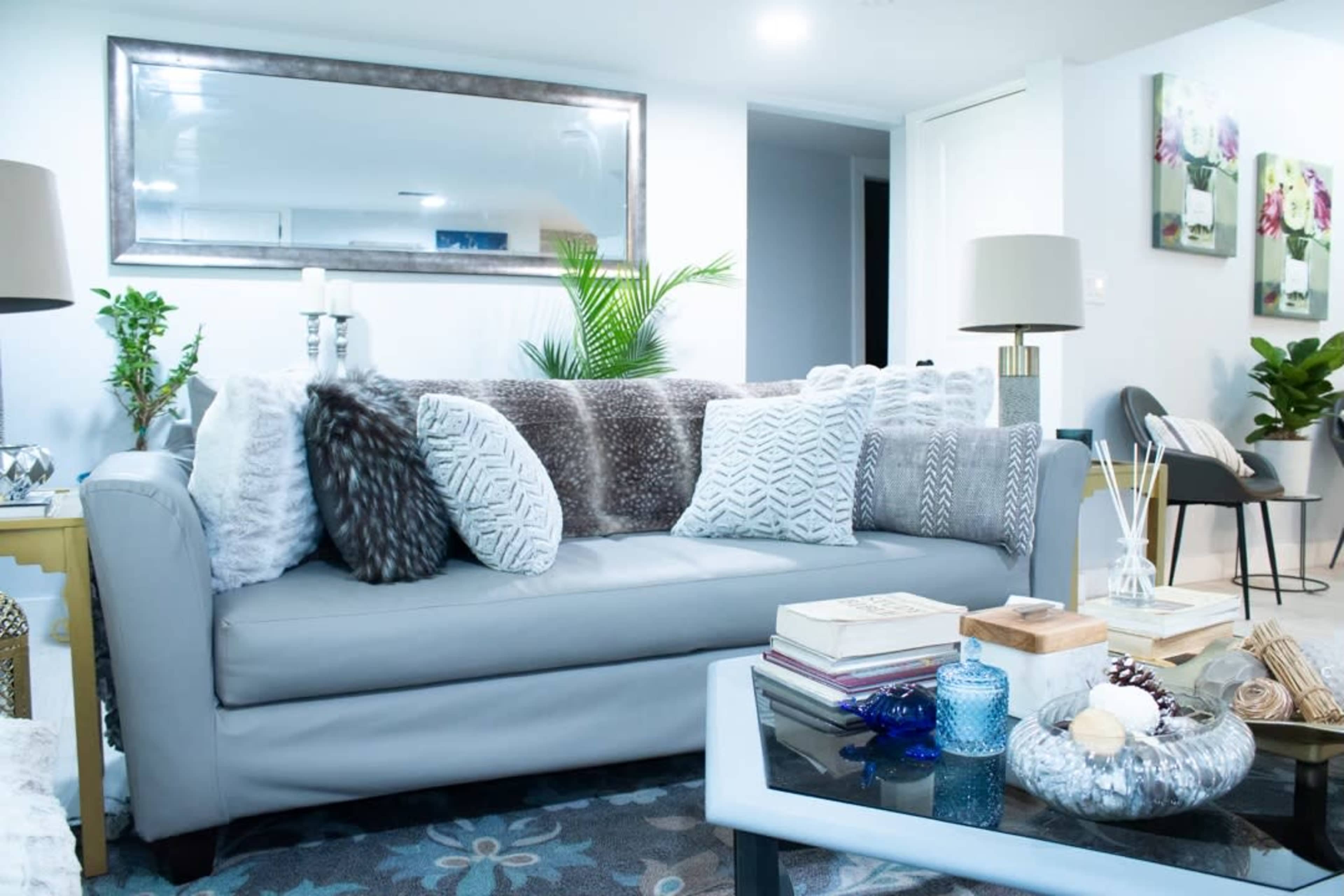 The image shows a modern living room featuring a gray sofa adorned with decorative pillows, a coffee table with books and decorative items, and walls decorated with framed artwork.