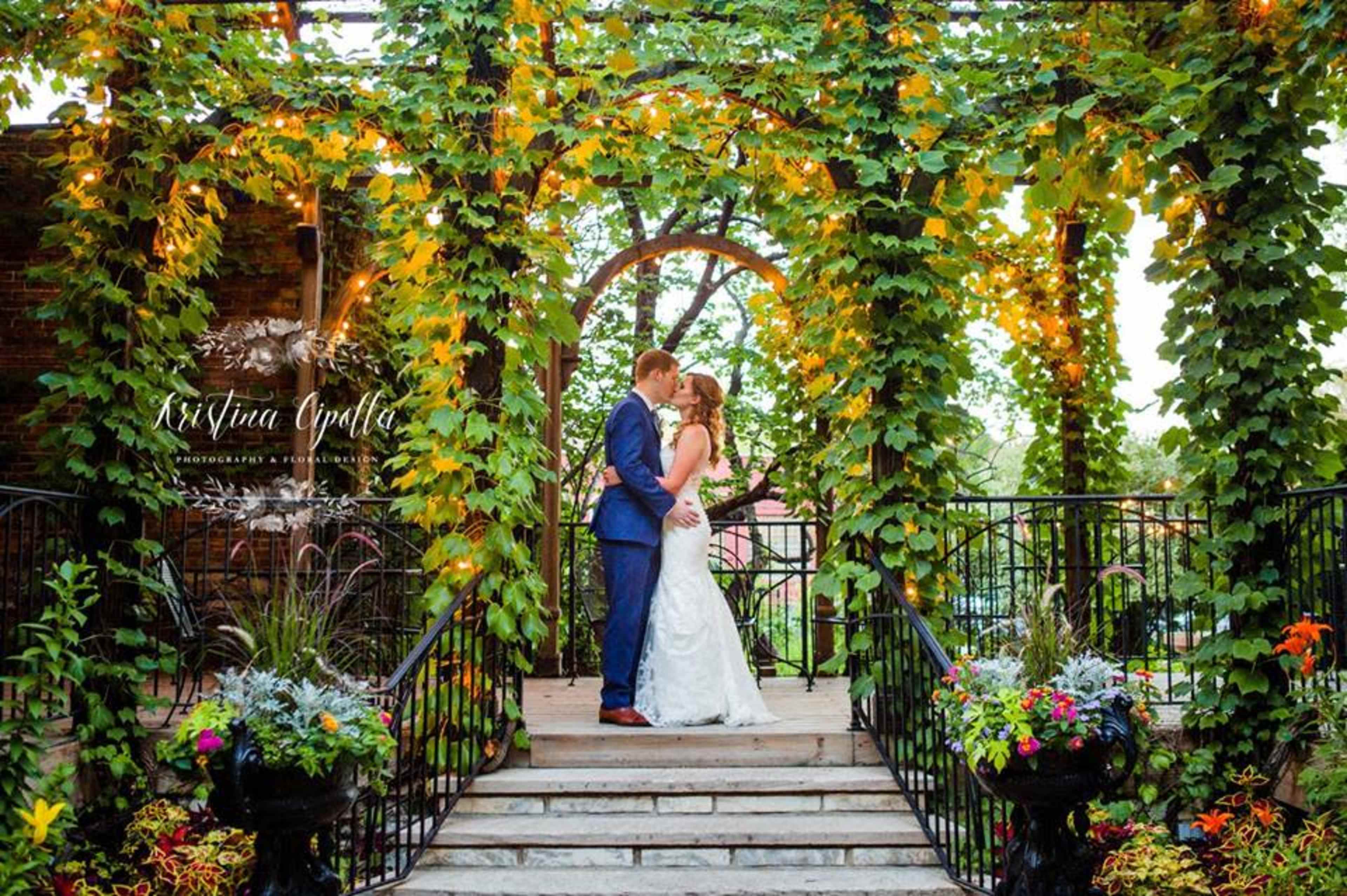 A couple stands on a wooden platform surrounded by greenery and decorative lights in a garden setting.