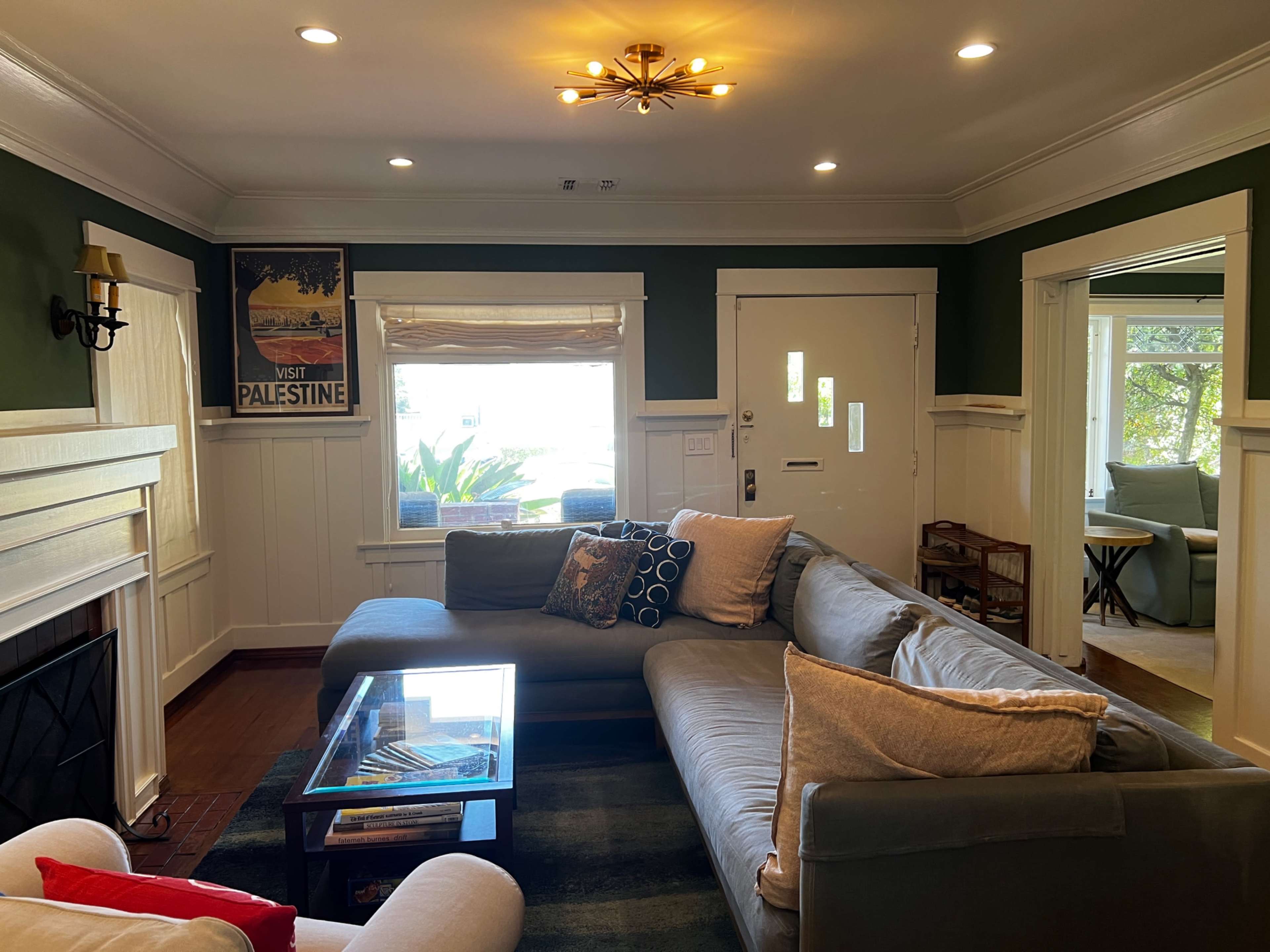 The image shows a cozy living room with a sectional couch, a coffee table, and a decorative wall poster, all surrounded by green-painted walls and natural light coming through the windows.