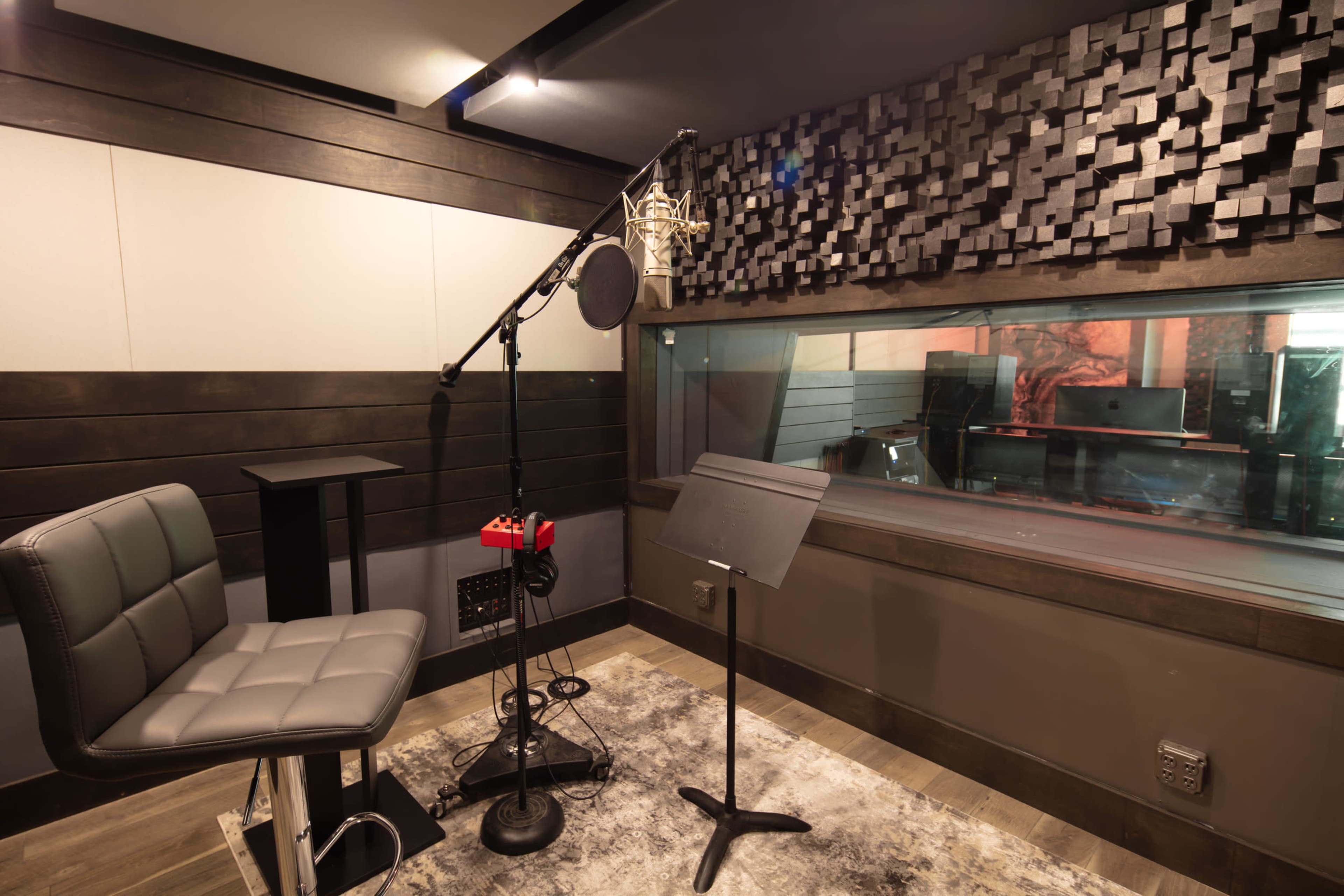 The image shows a soundproof recording studio featuring a microphone on a stand, a chair, and a music stand in front of a glass window.