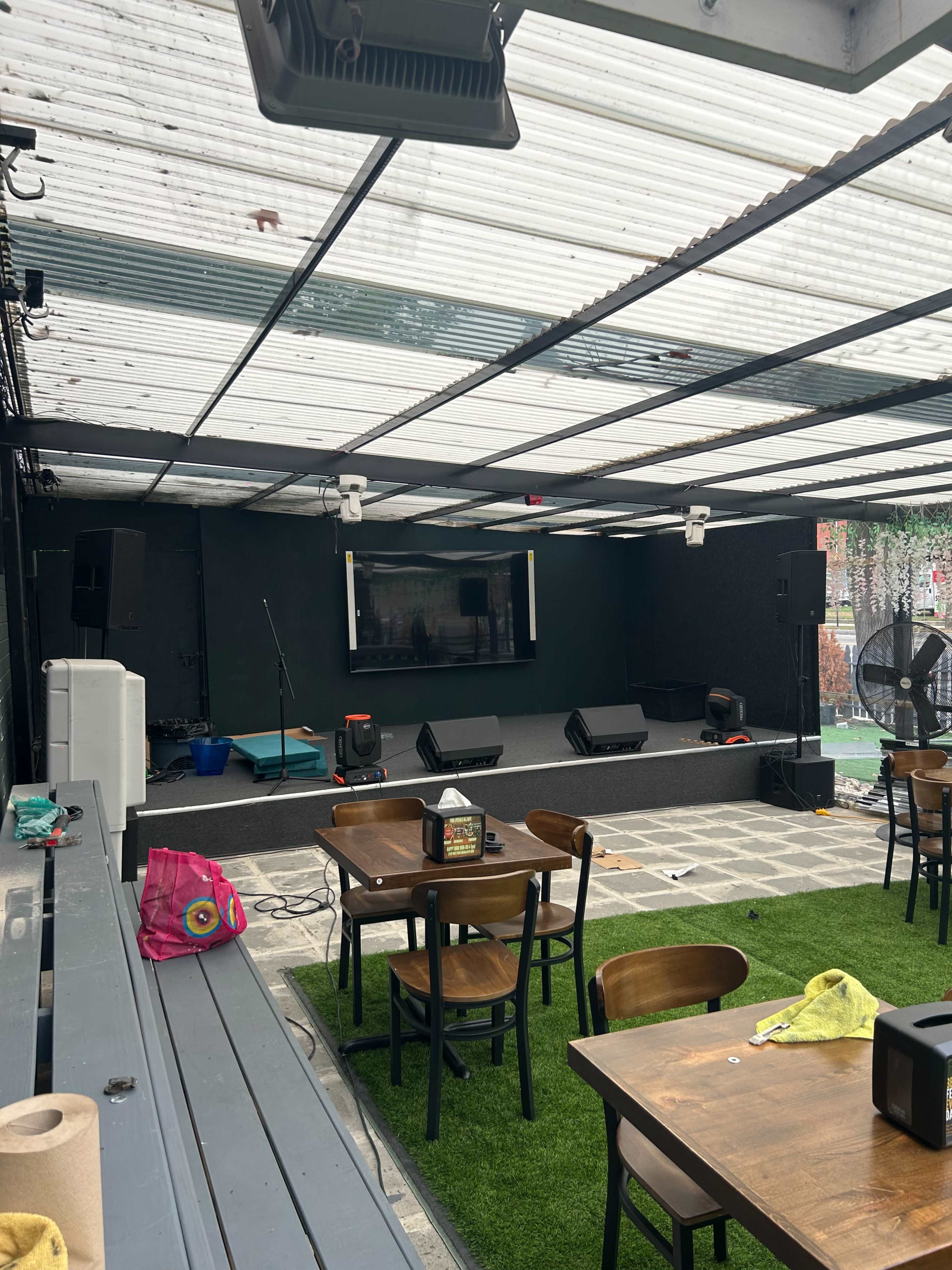 An outdoor stage setup with a sound system, tables, and chairs arranged under a covered area.