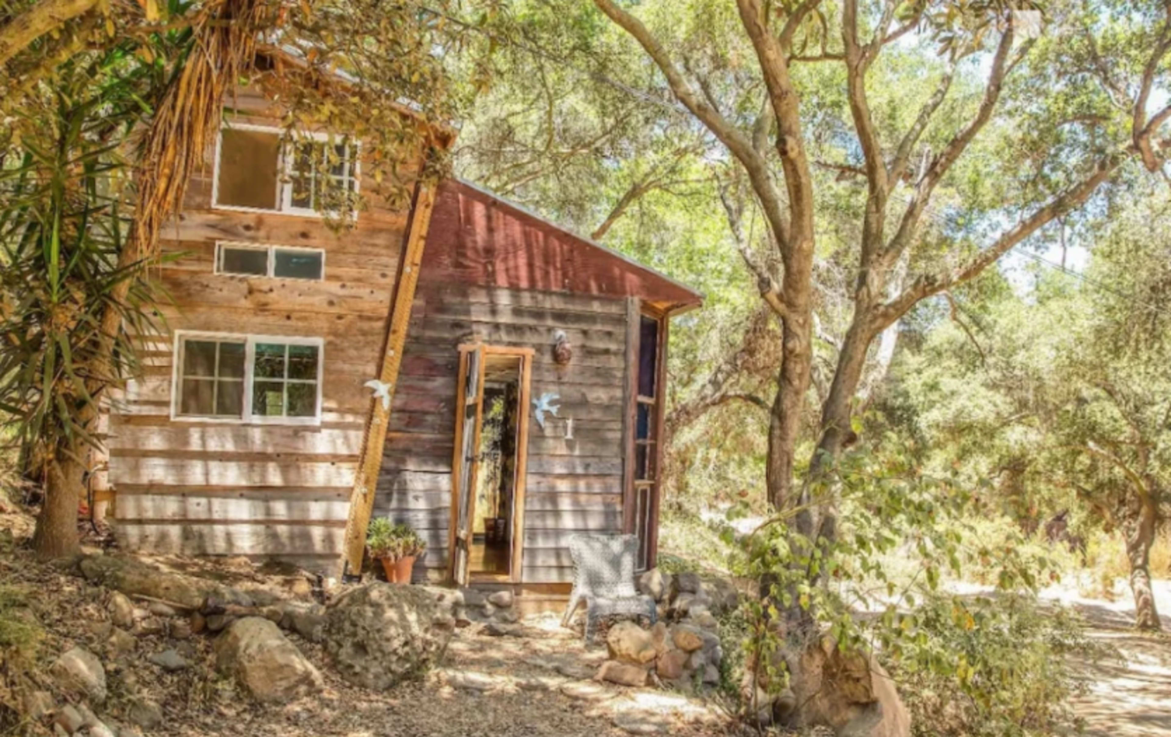 A small wooden cabin with a slanted roof is situated among trees and rocks in a natural setting.