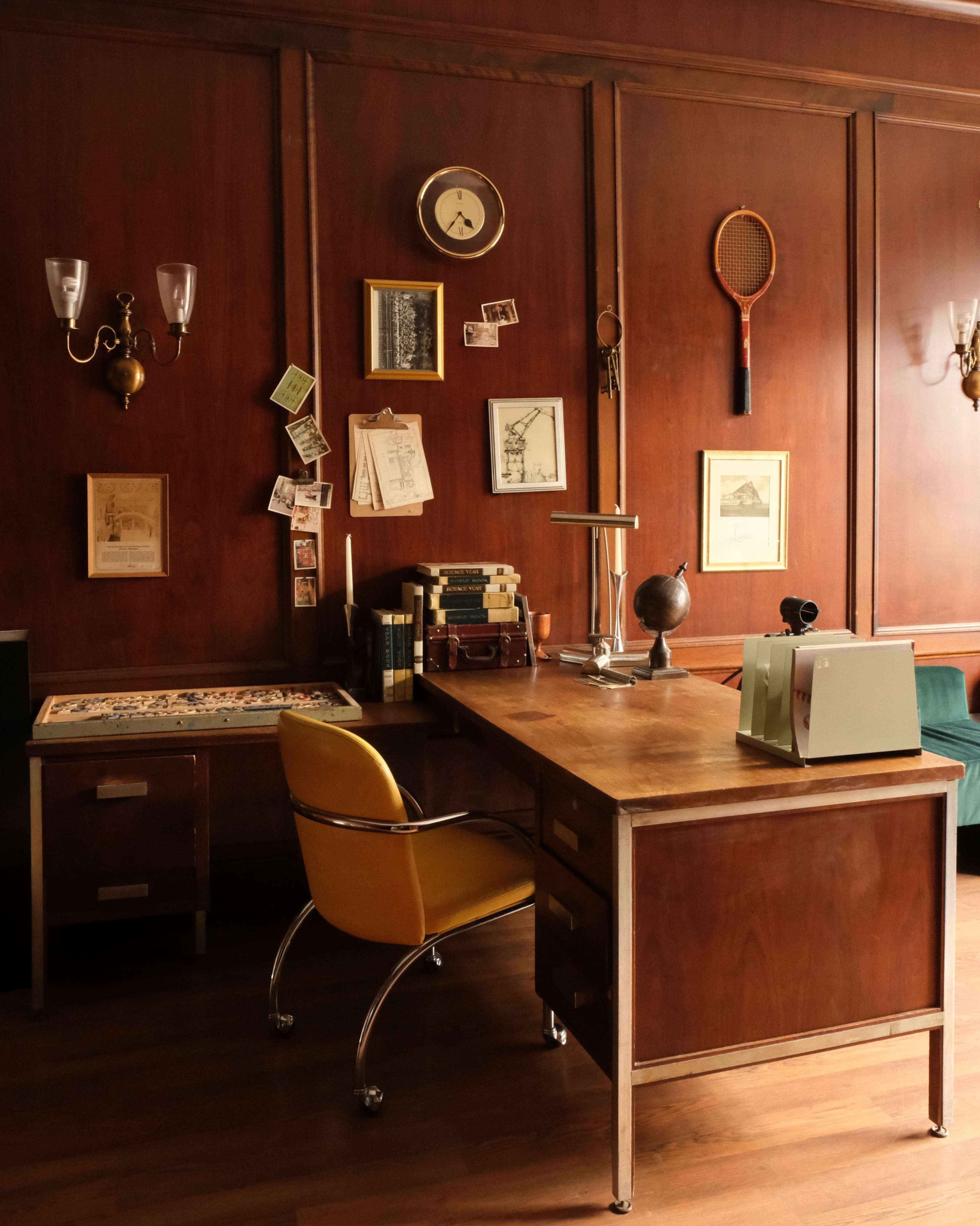 The image shows a wooden office space with a desk, a yellow swivel chair, a globe, and various framed pictures and decor mounted on the wall.