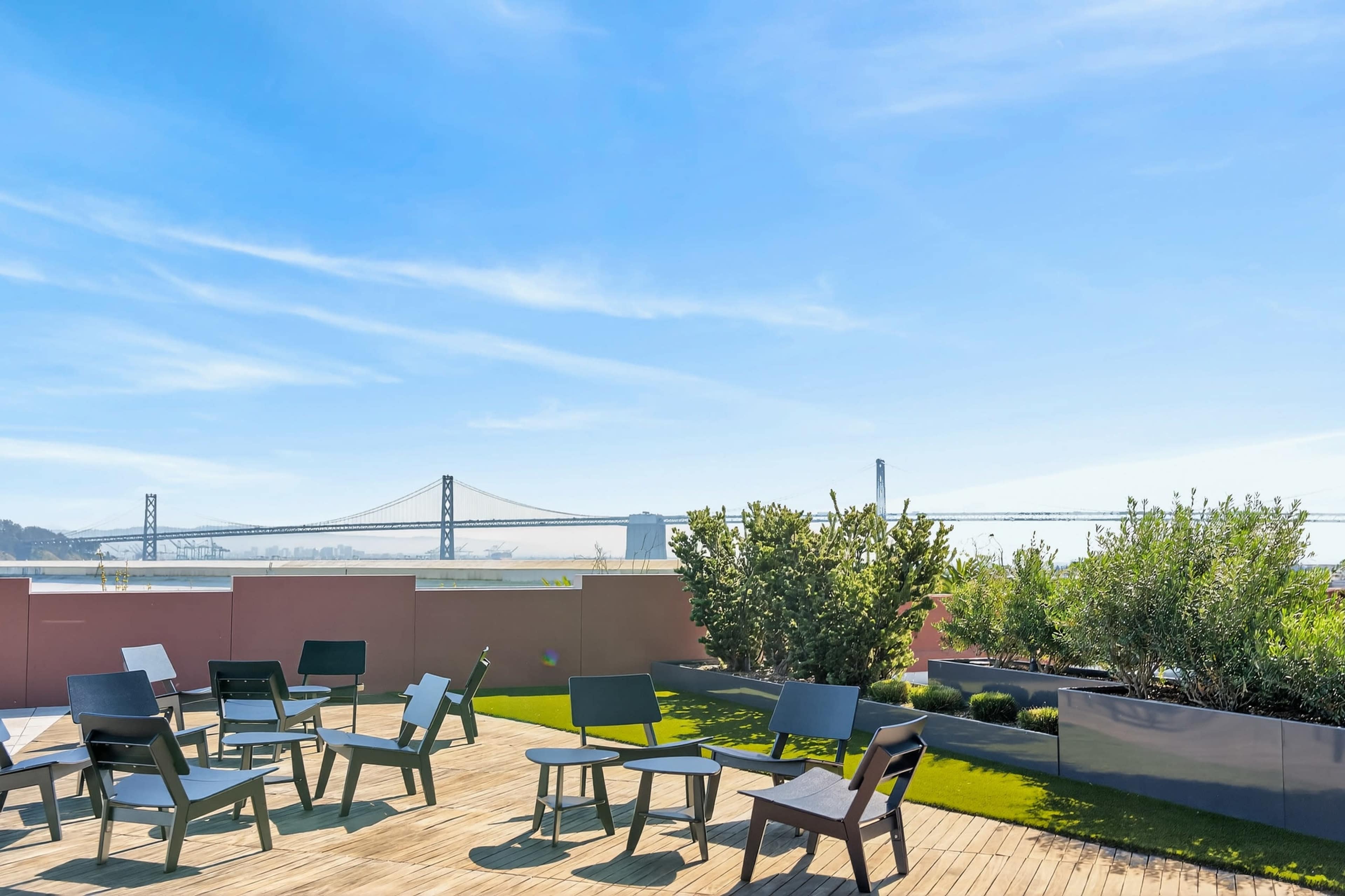 The image shows a rooftop deck with several chairs arranged around a low wooden table, overlooking a bay and a bridge in the background.