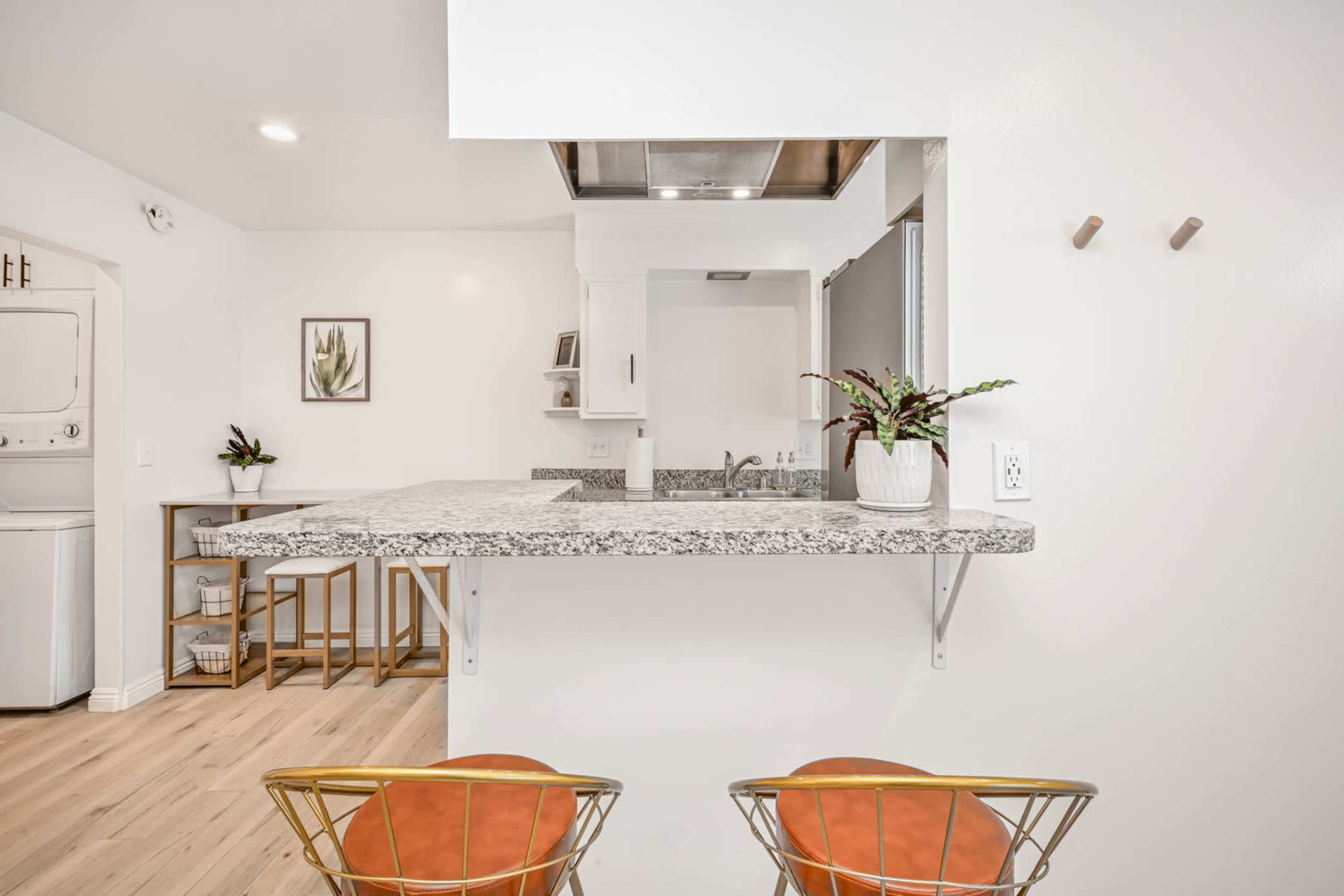 A modern kitchen features a granite countertop with two metal chairs, adjacent to a laundry area and a small dining space.