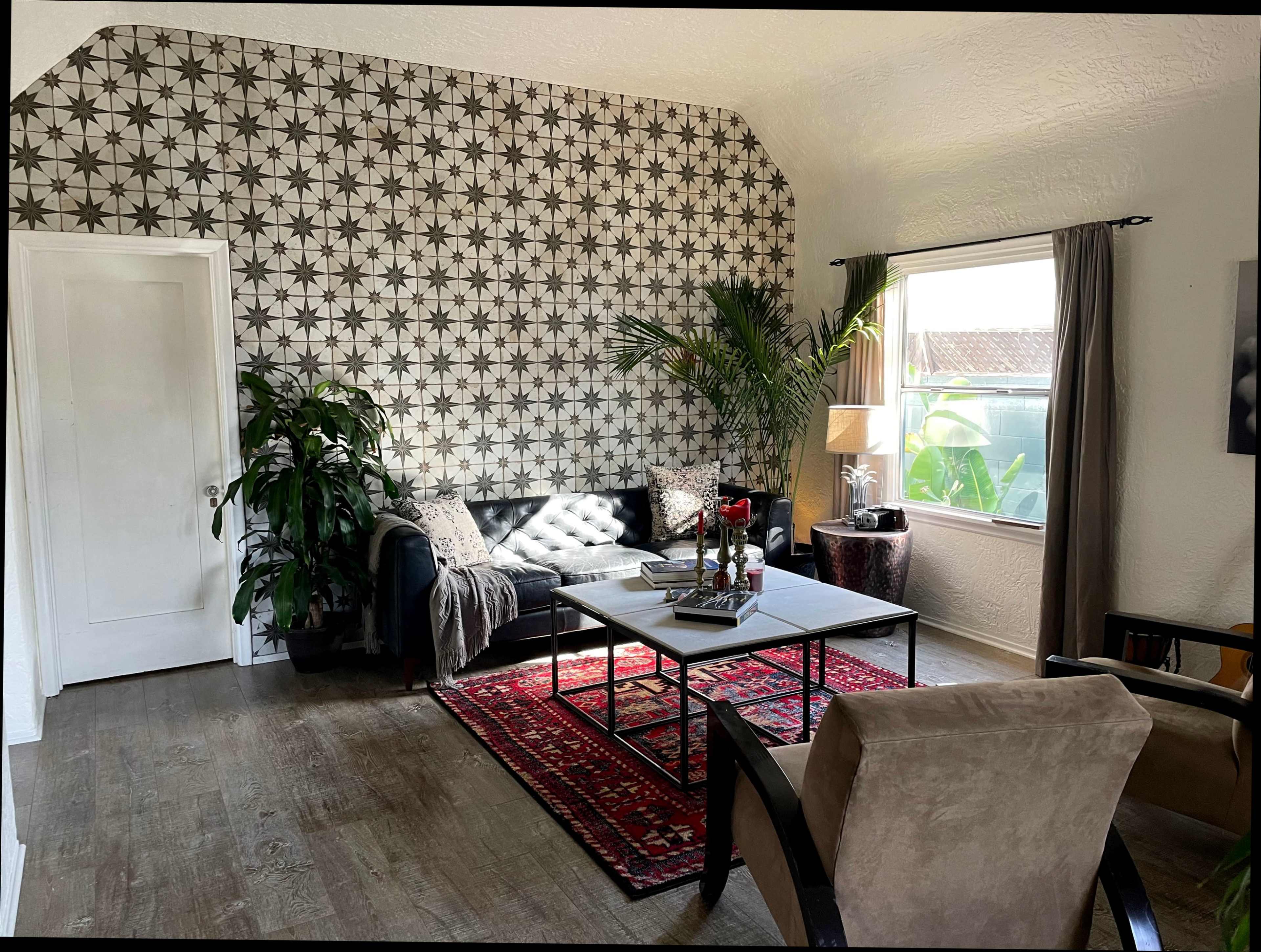 The image shows a living room with a patterned wallpaper, a black sofa, a coffee table, and two armchairs, along with plants and a window providing natural light.