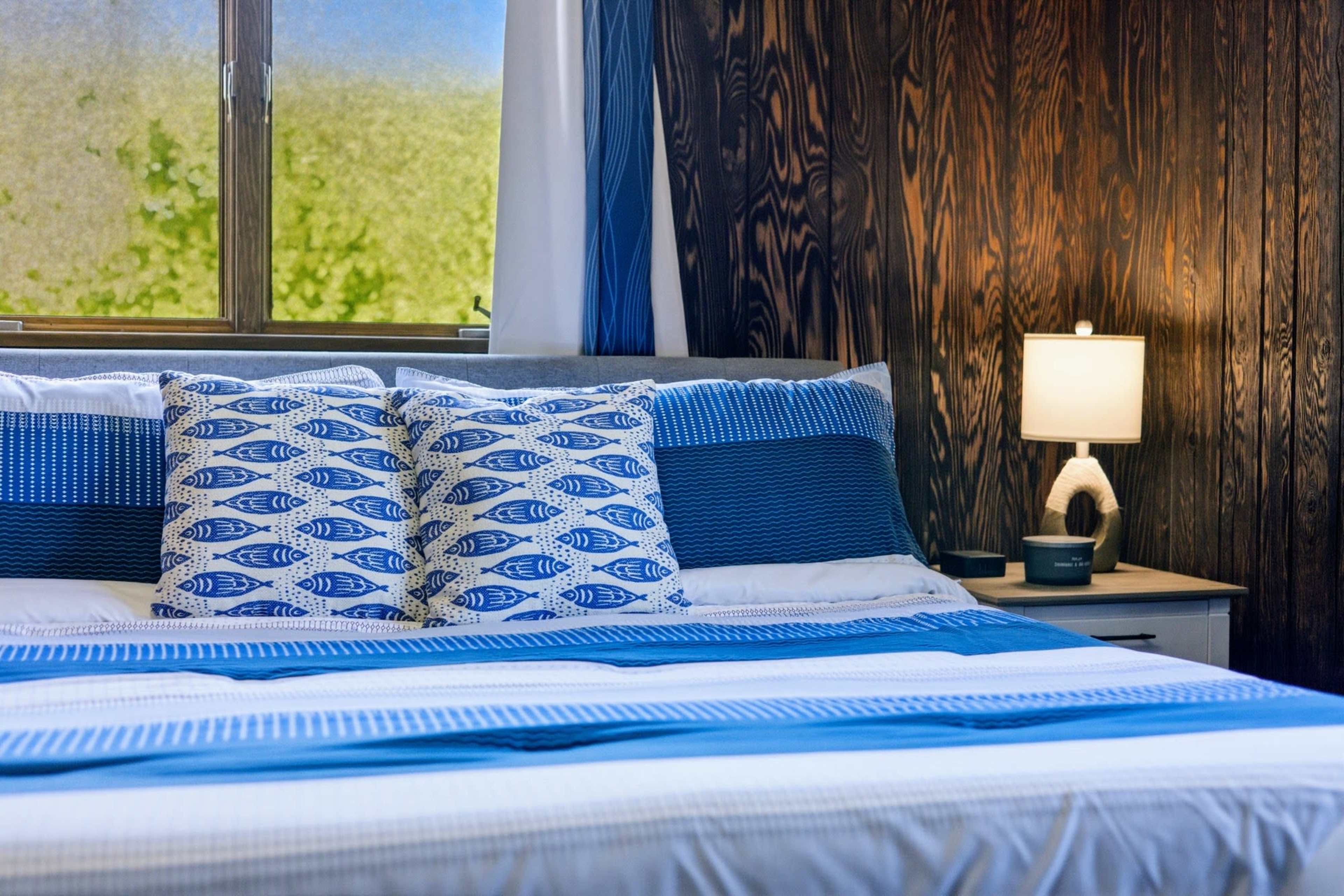 A neatly made bed with blue and white striped bedding and decorative pillows is positioned next to a nightstand with a lamp in a room featuring dark wooden walls.