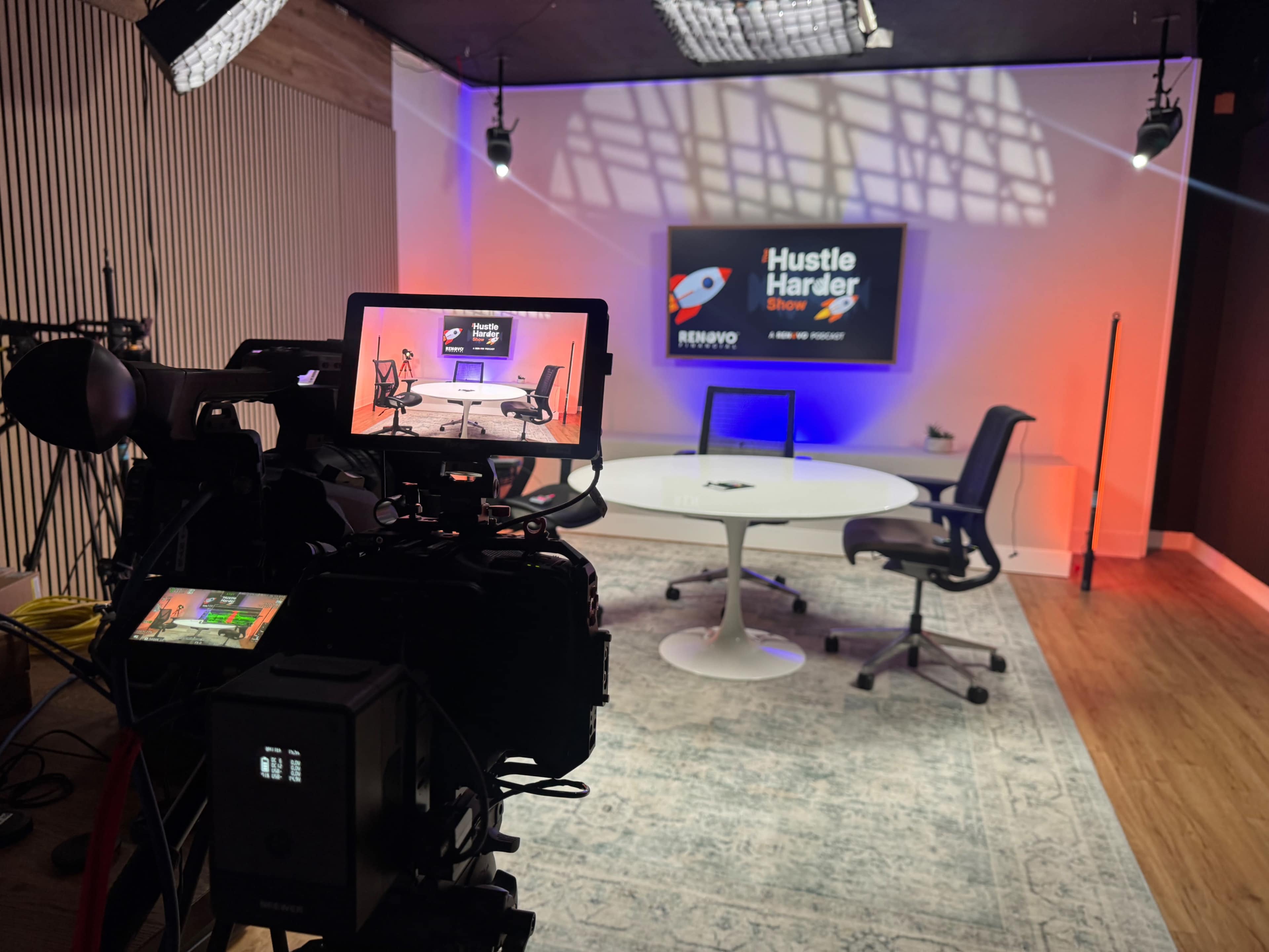 The image shows a well-lit studio setup with a camera pointed towards a round table and chairs, featuring a screen displaying "Hustle Harder Show."
