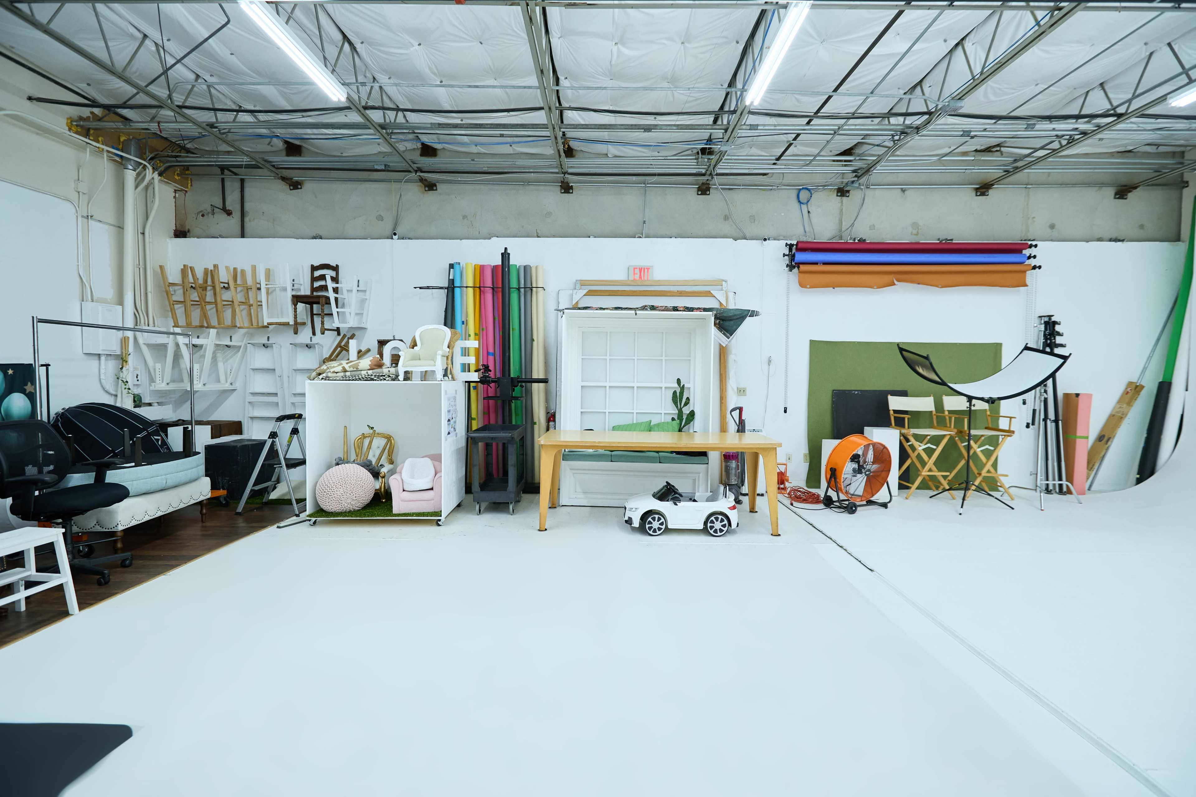 A spacious photography studio with various props, backdrops, and furniture arranged on a white floor.