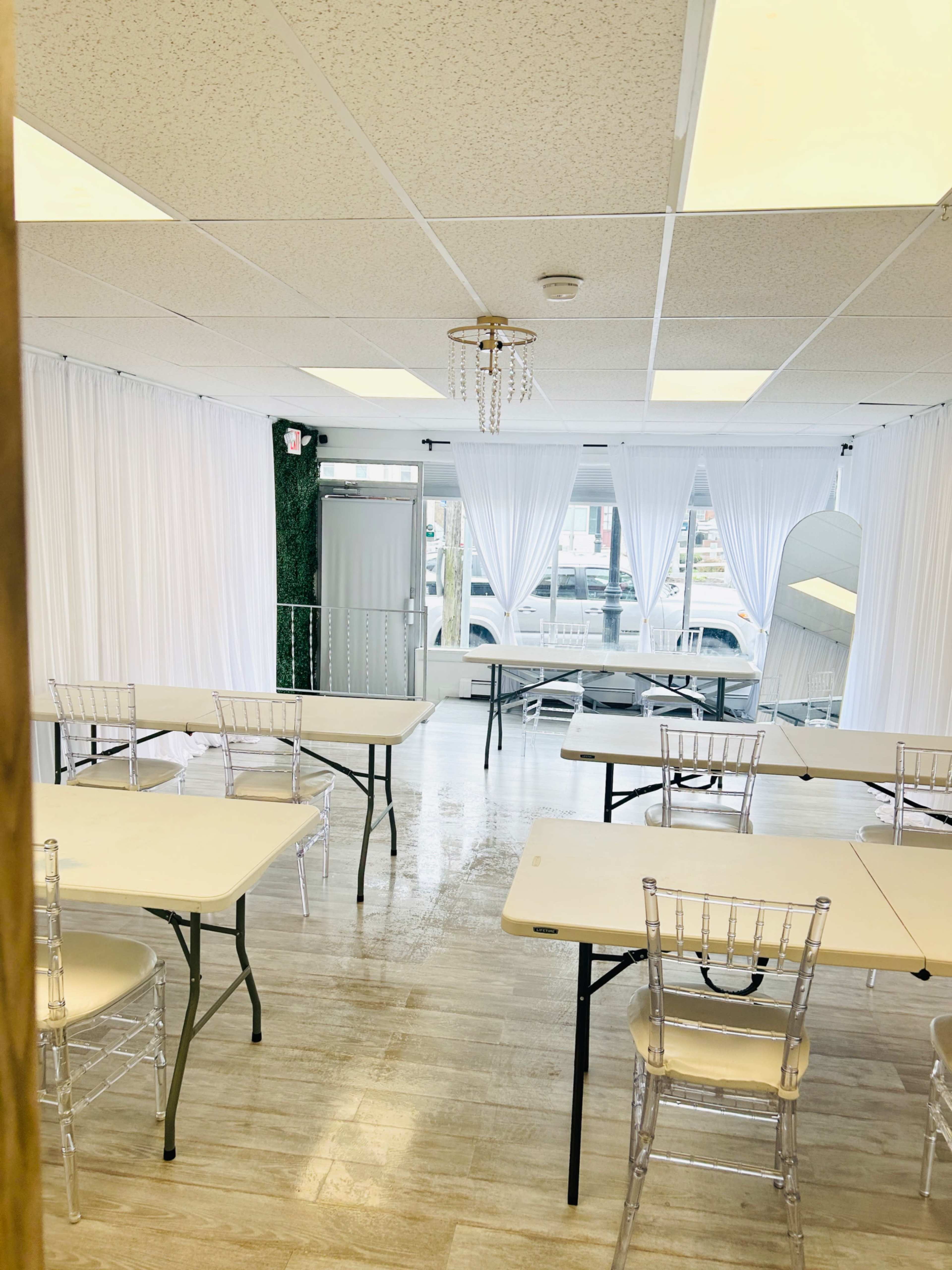 The image shows a bright event space with multiple tables and chairs arranged neatly, white drapes hanging on large windows, and clean, polished flooring.
