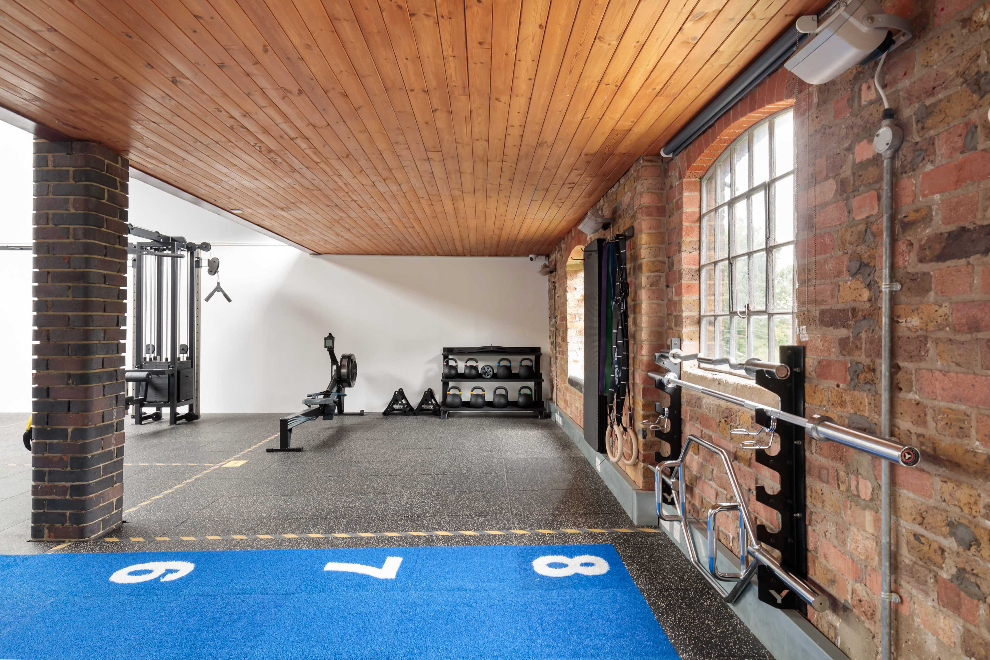 A spacious gym with a blue rubber floor, wooden ceiling, and various fitness equipment arranged against brick walls.