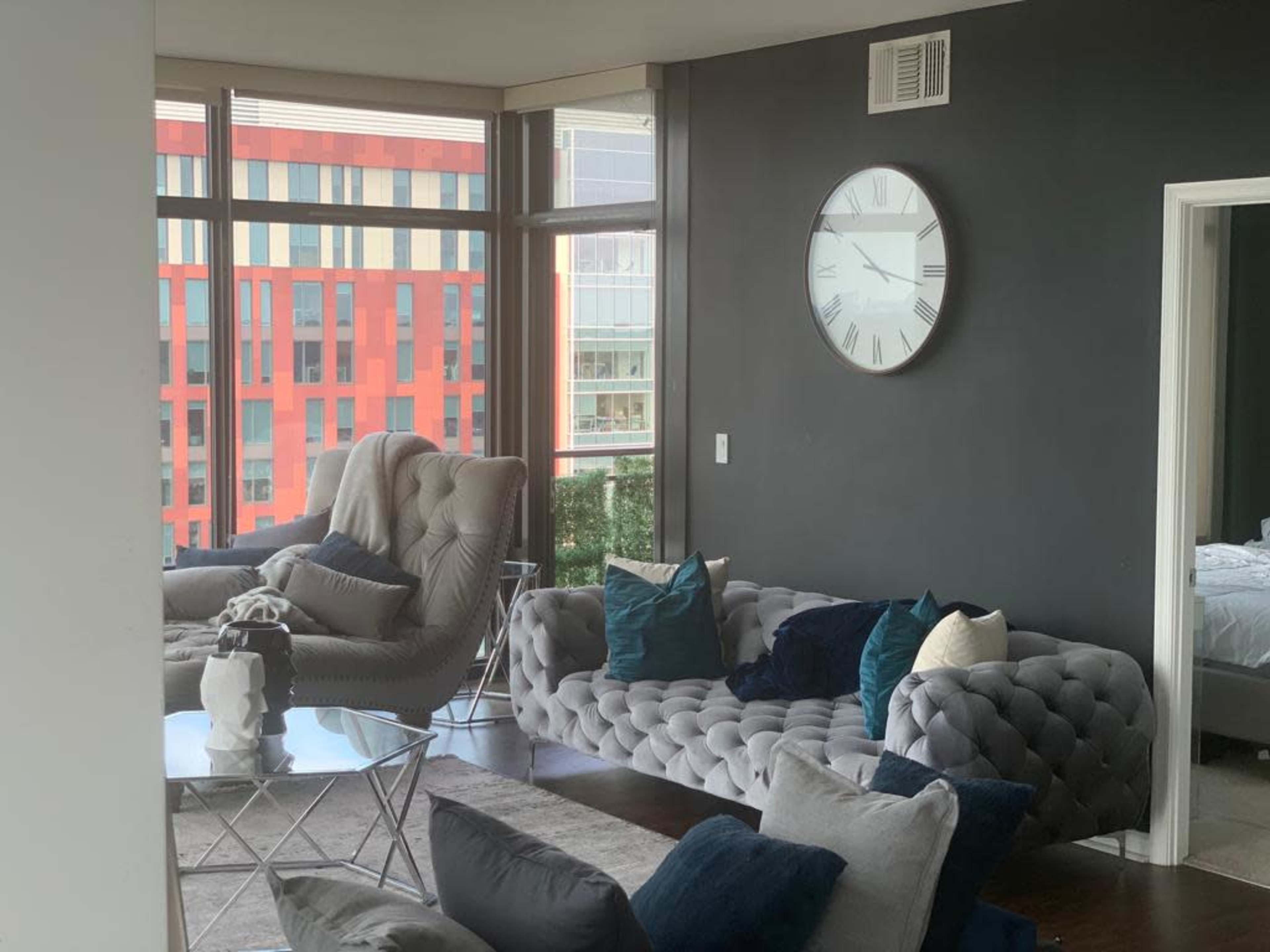 A modern living room with a gray sectional sofa, a glass coffee table, and large windows overlooking a colorful building.