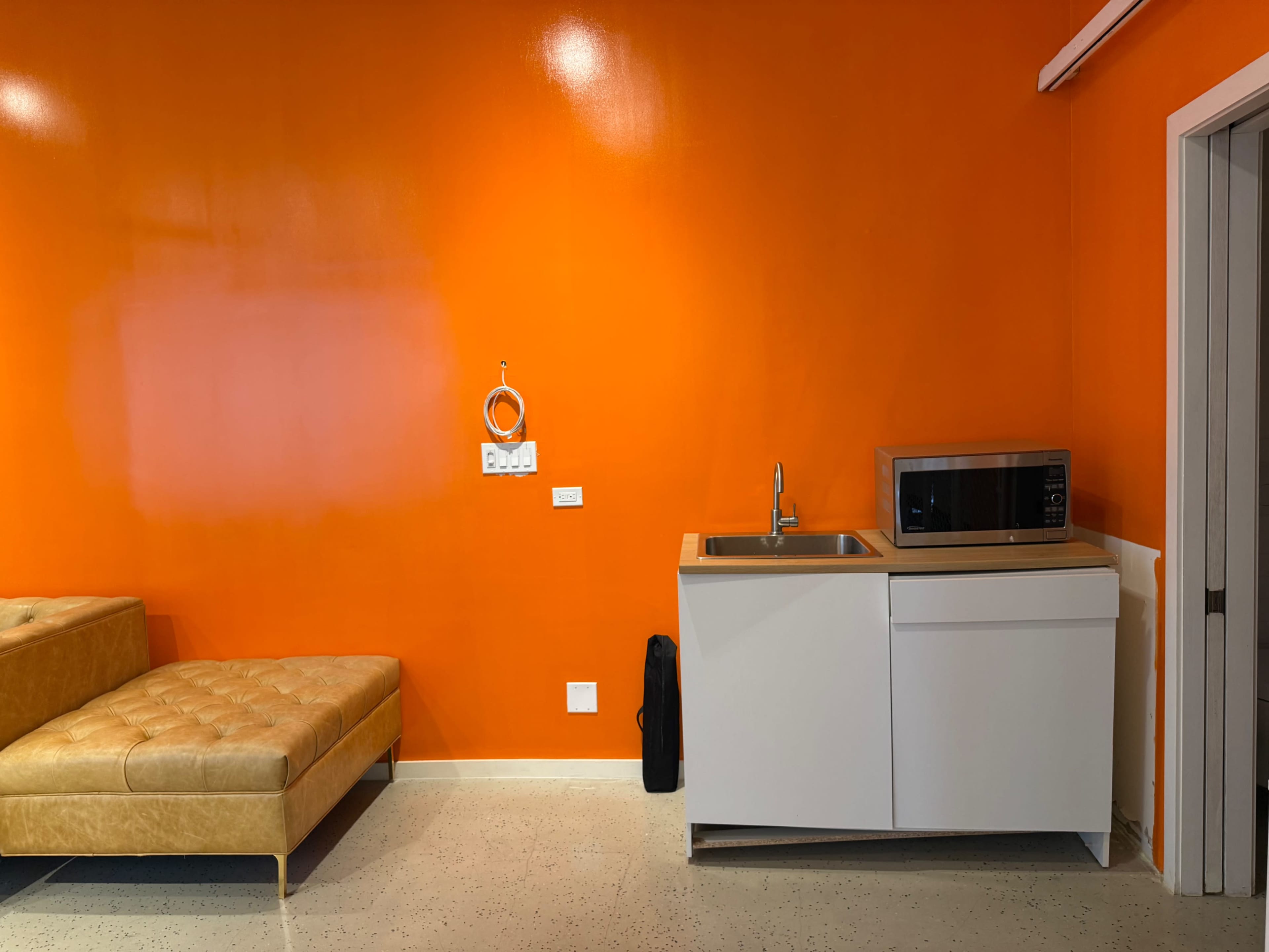 The image features a bright orange wall with a small kitchen area that includes a sink, a microwave, and a beige sofa nearby.