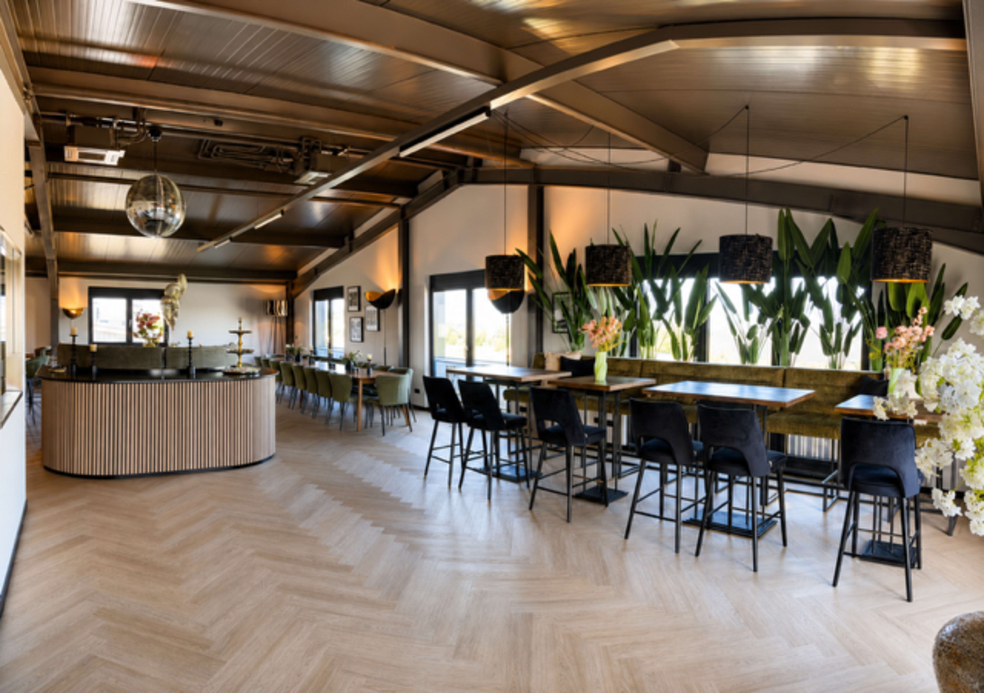 The image shows a spacious dining area with a bar counter, tall tables, and large windows surrounded by decorative plants.