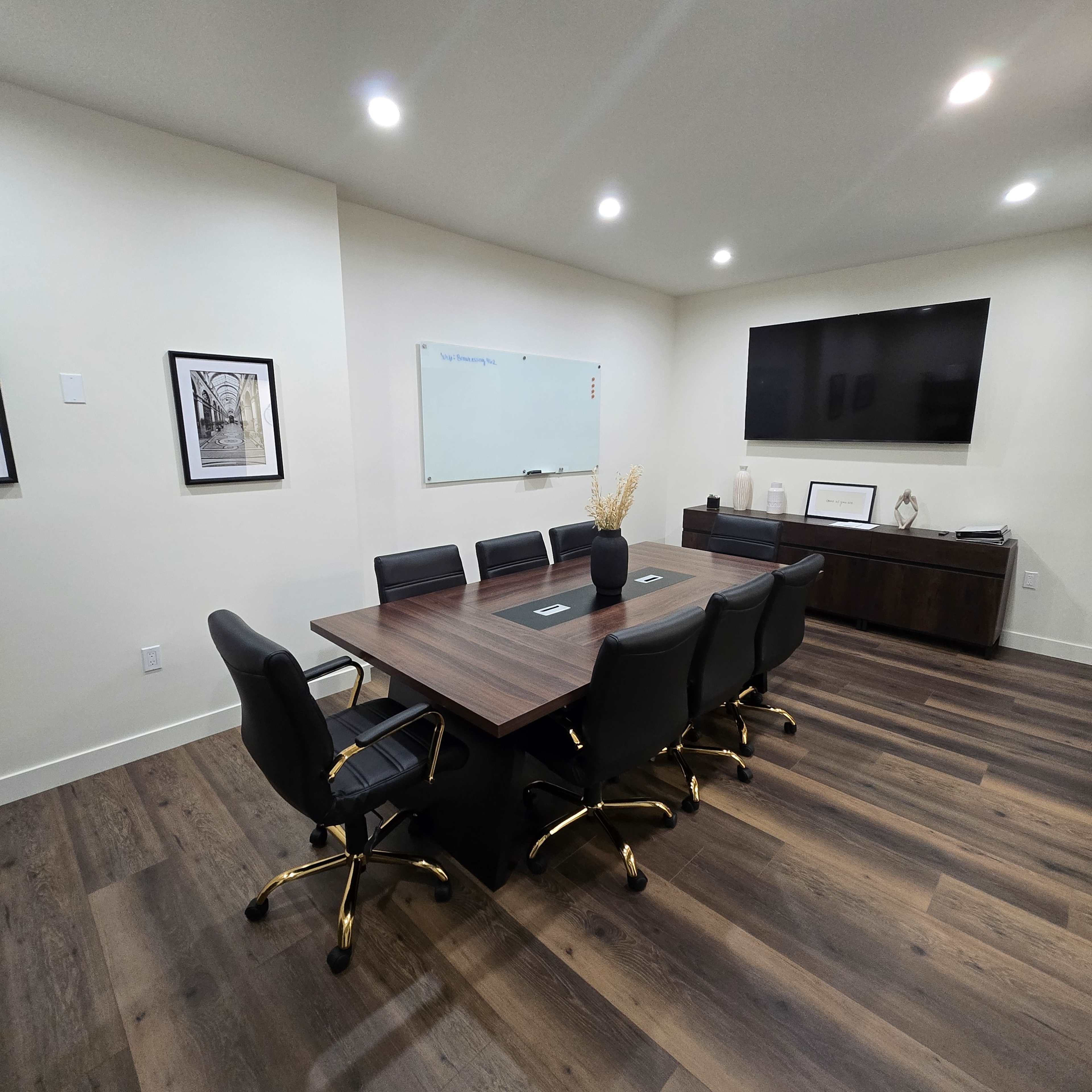 The image shows a modern conference room with a wooden table surrounded by black chairs, a wall-mounted TV, a whiteboard, and framed artwork.