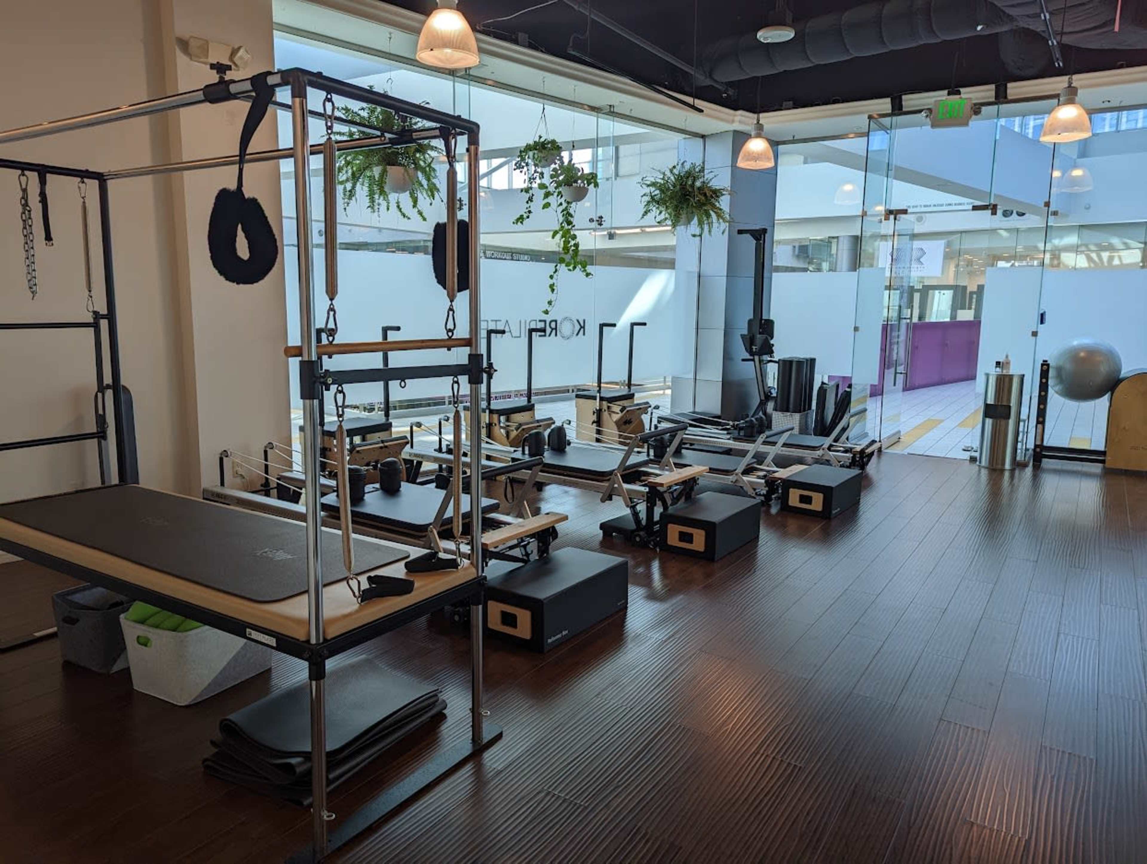 The image shows a brightly lit fitness studio featuring various Pilates equipment, including reformers, a reformer barrel, and mats arranged neatly on the wooden floor.