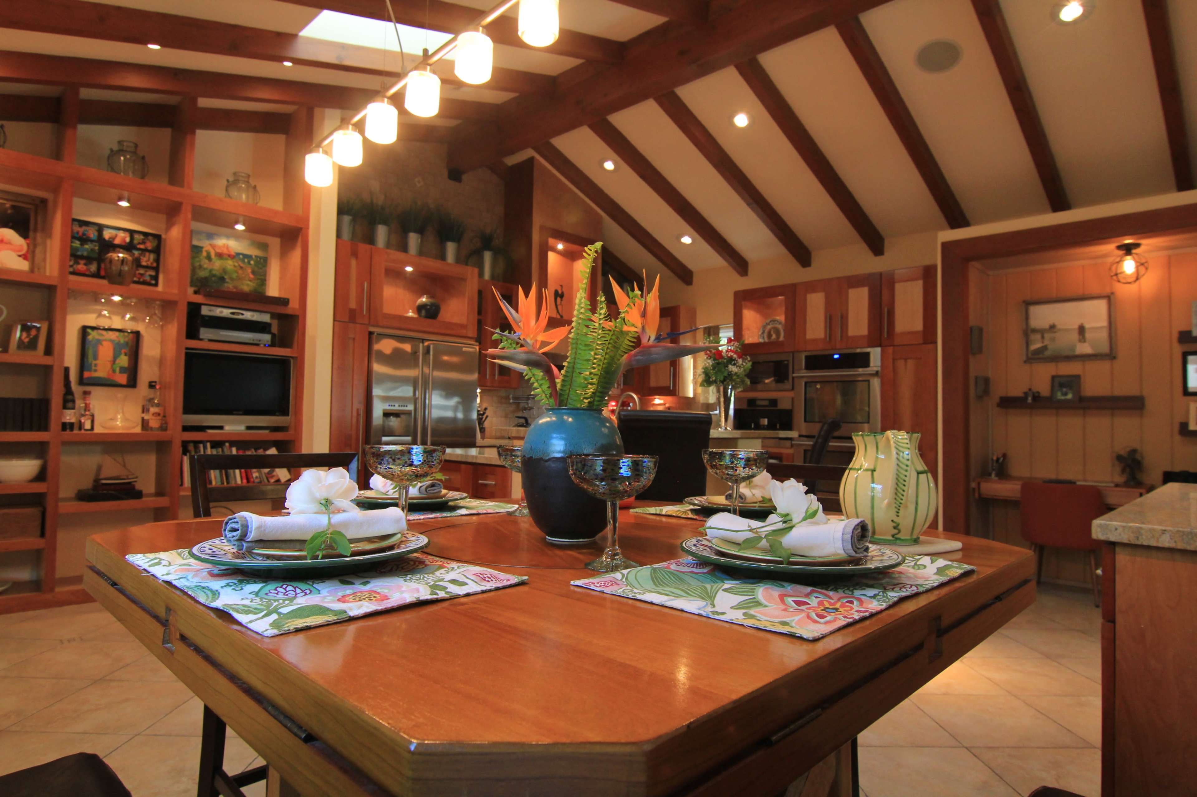 A dining table is set with plates and flowers in a bright kitchen featuring wooden beams and built-in shelves.