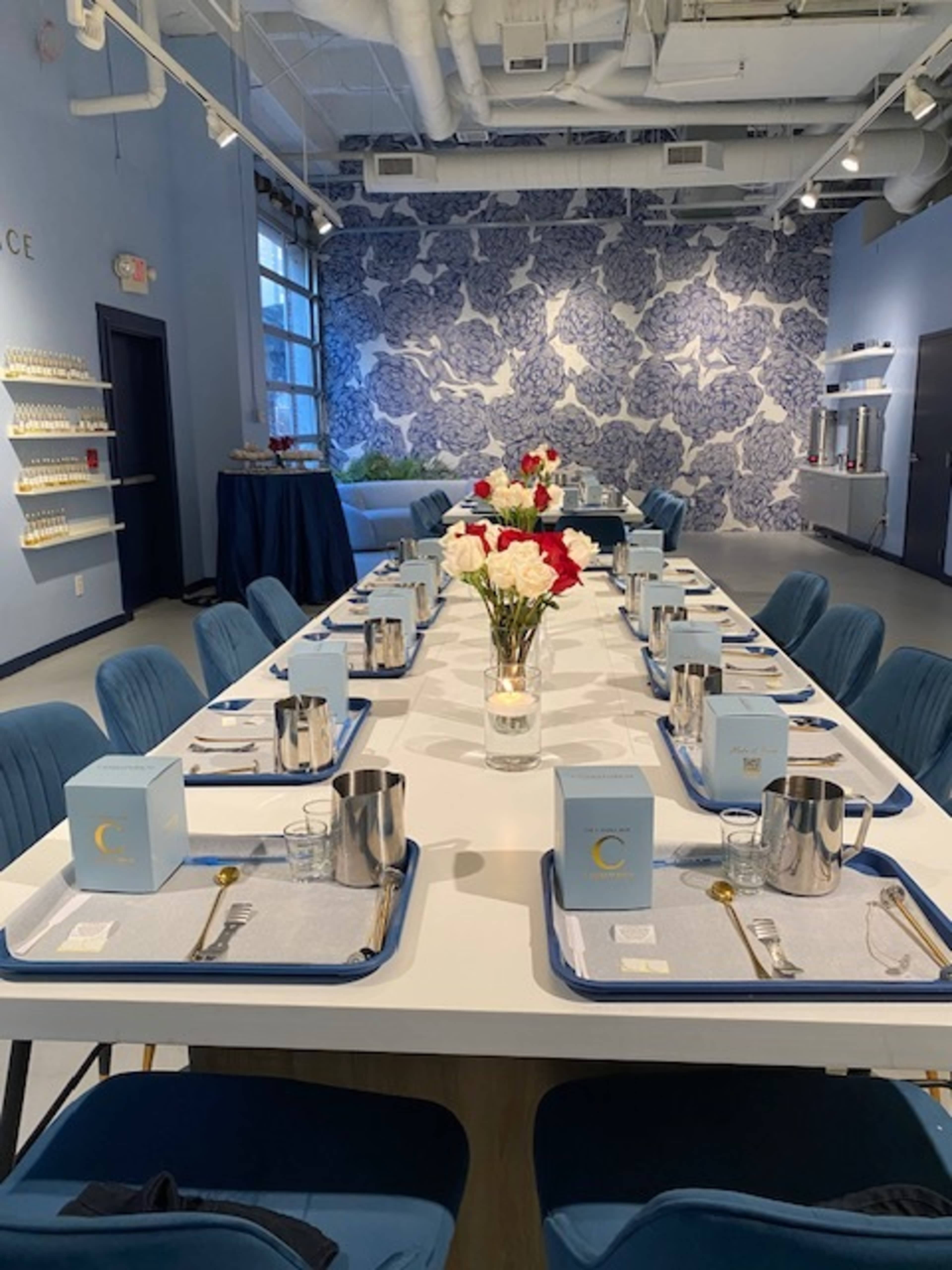 A long dining table is set with blue trays, glasses, and light blue boxes, surrounded by plush chairs, against a backdrop of patterned wallpaper and floral decorations.