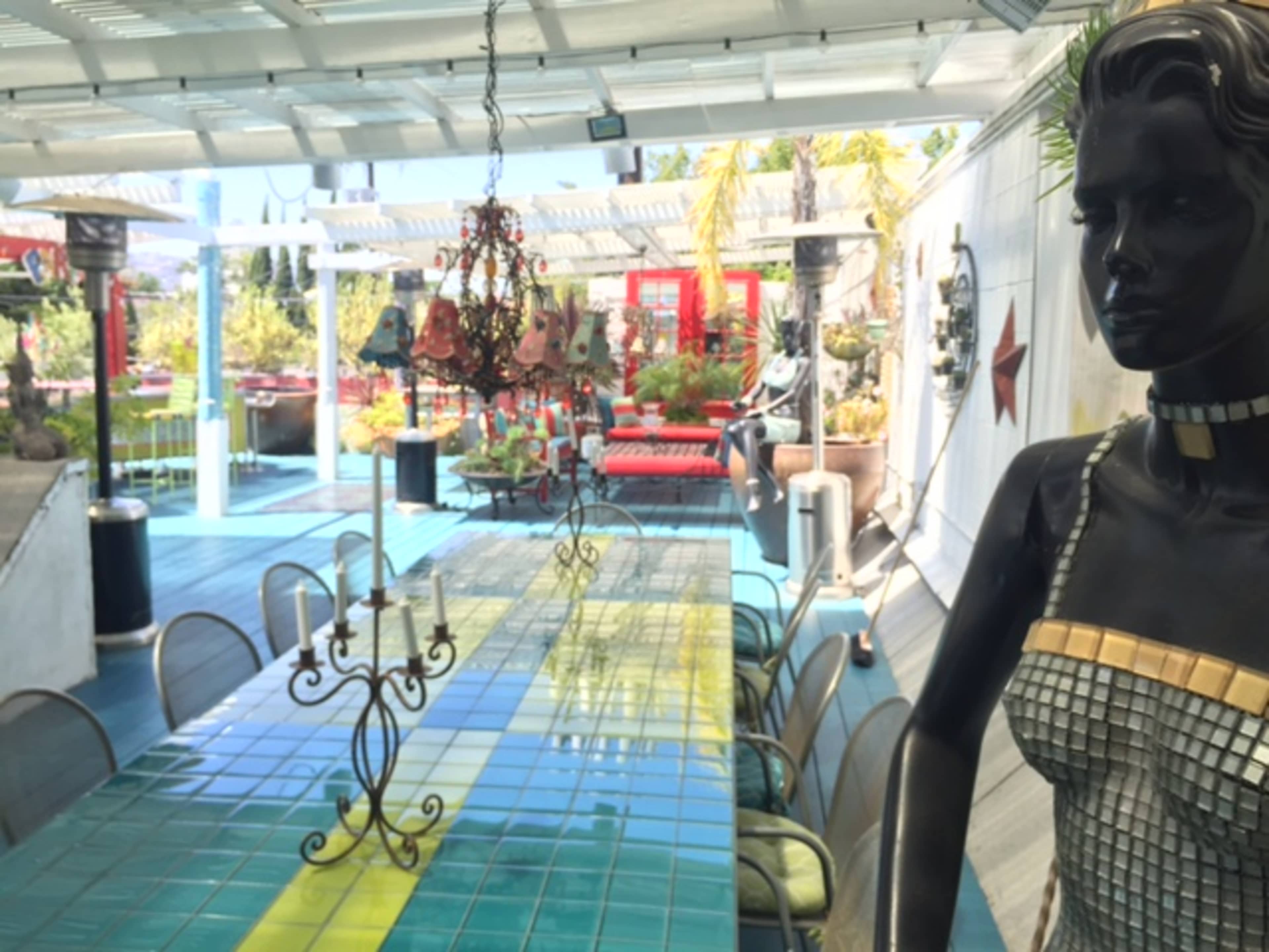 A long, colorful table is set under a covered patio, accompanied by a black statue of a woman and decorative plants in the background.