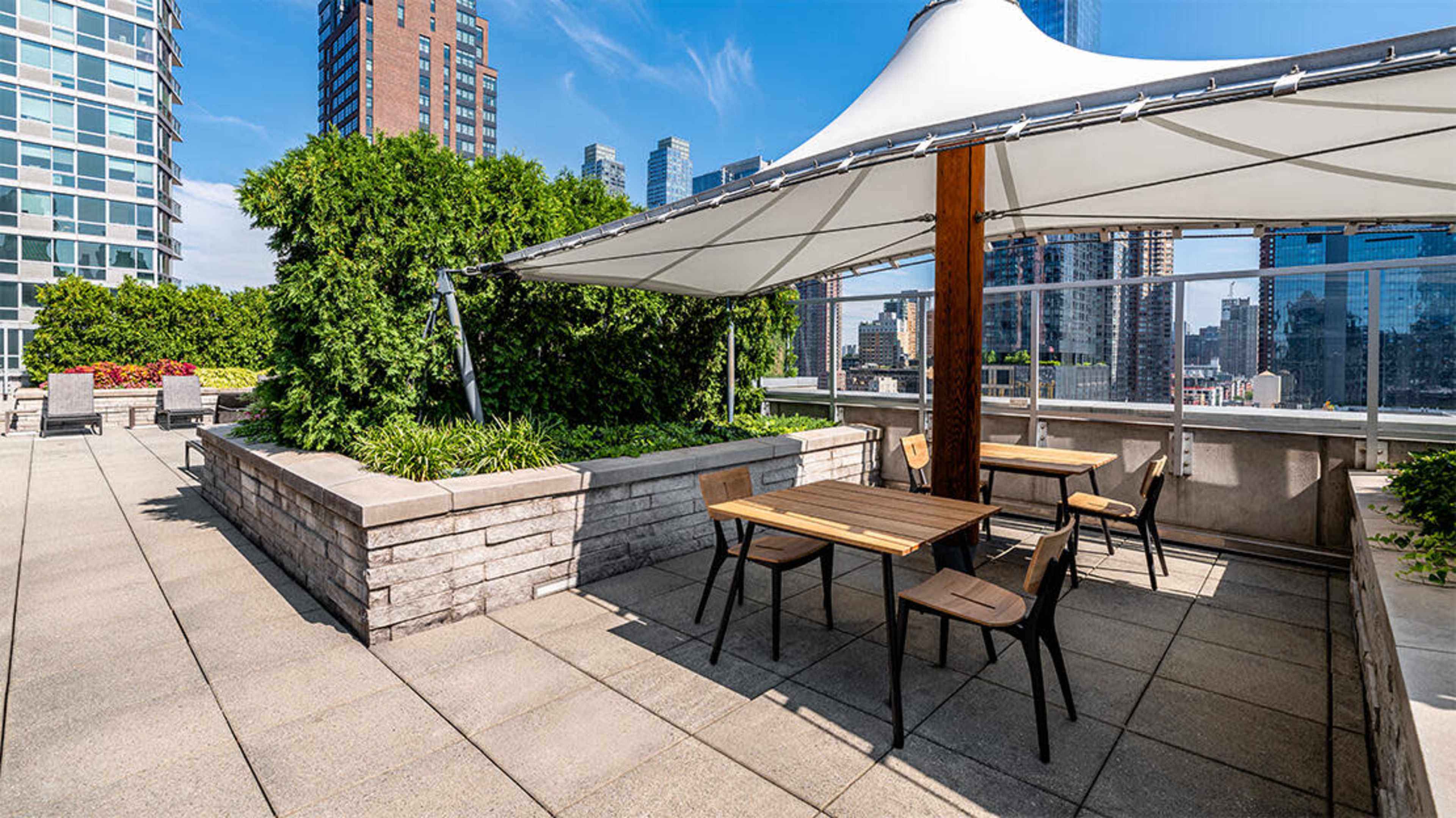 A rooftop terrace features a shaded area with wooden tables and chairs, surrounded by greenery and city skyscrapers in the background.