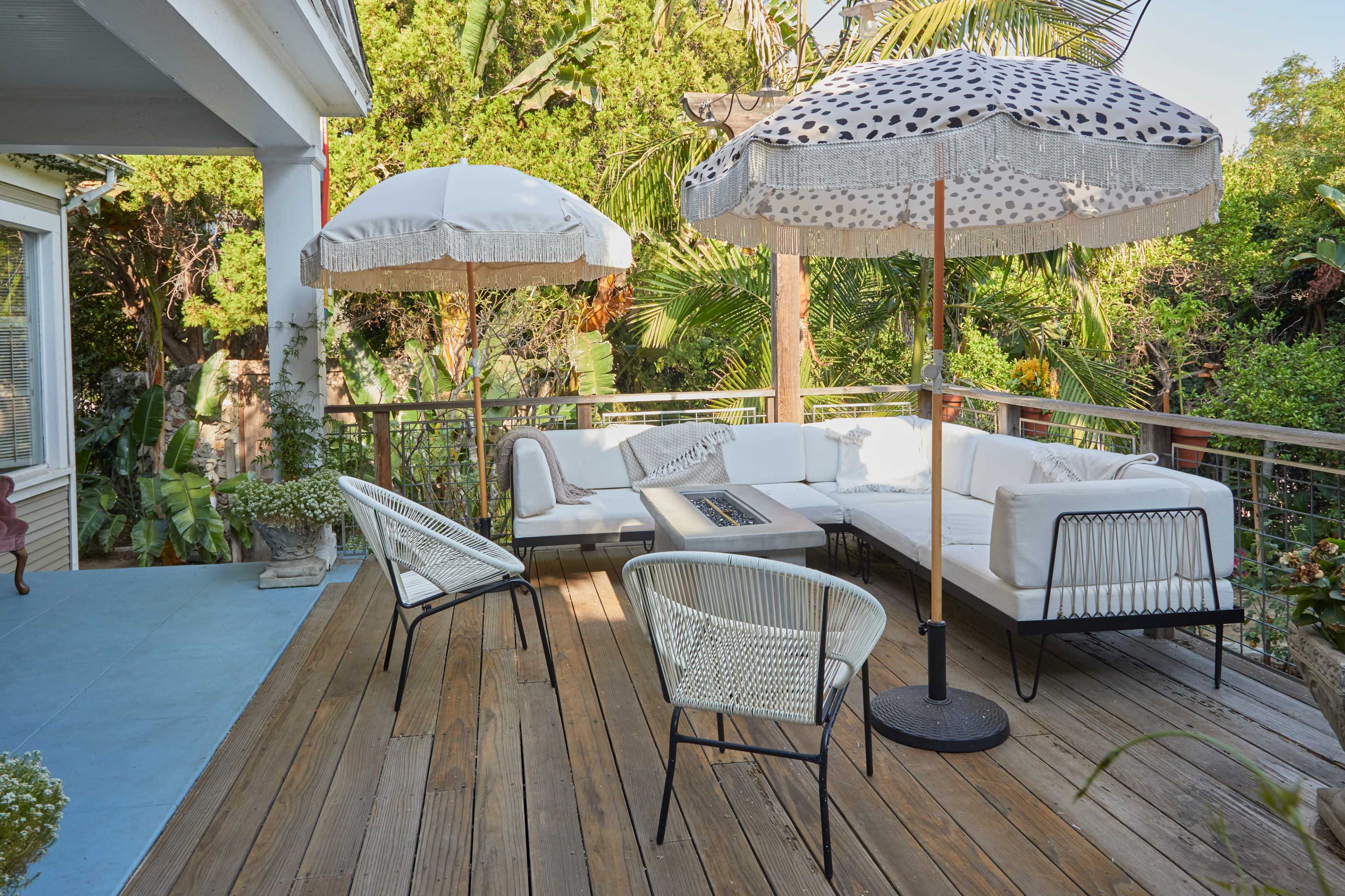 A deck features a sectional sofa and four chairs, shaded by two large umbrellas surrounded by lush greenery.
