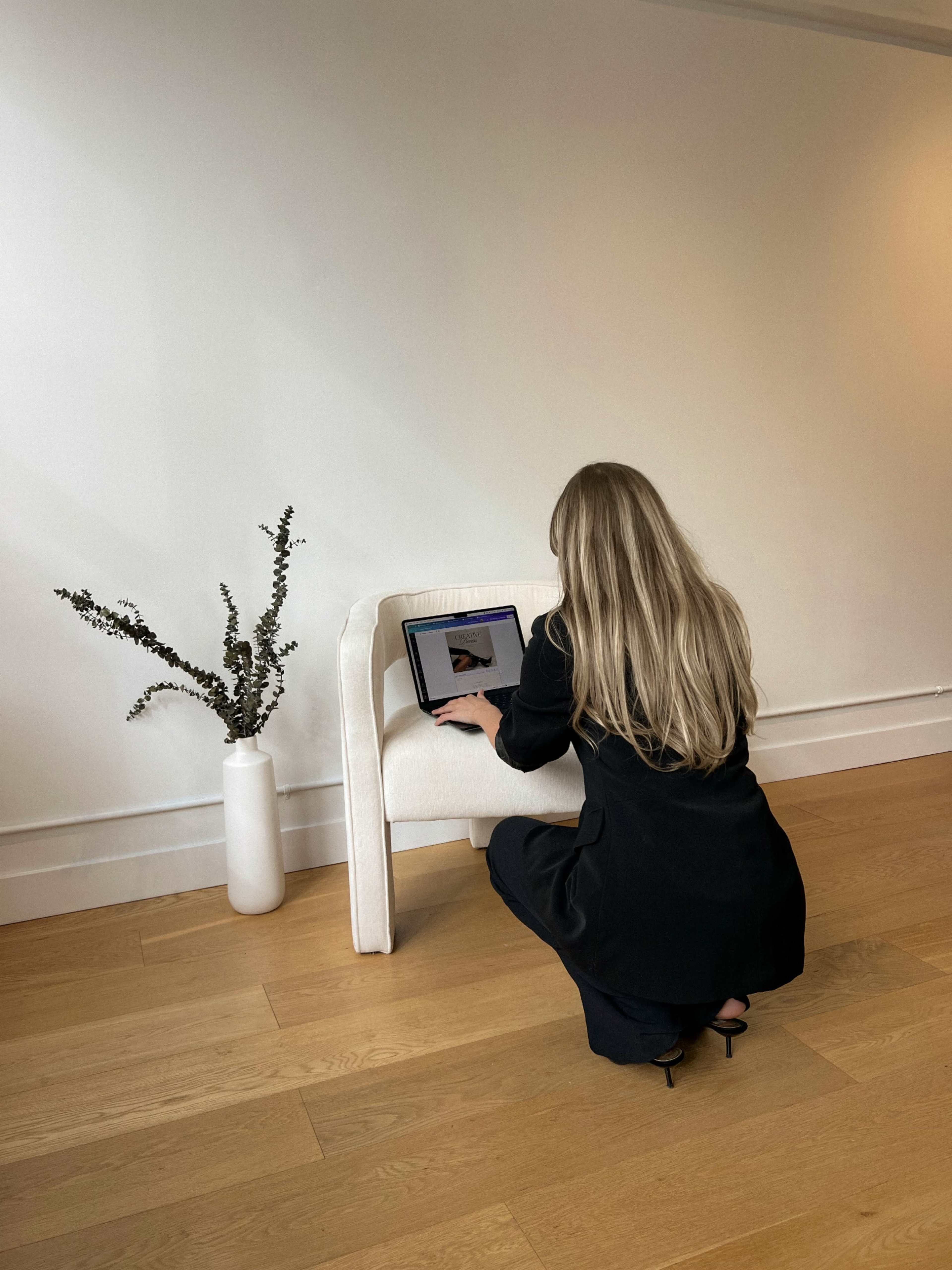 A woman with long hair is kneeling beside a white chair, working on a laptop placed on it, with a vase of greenery in the background.