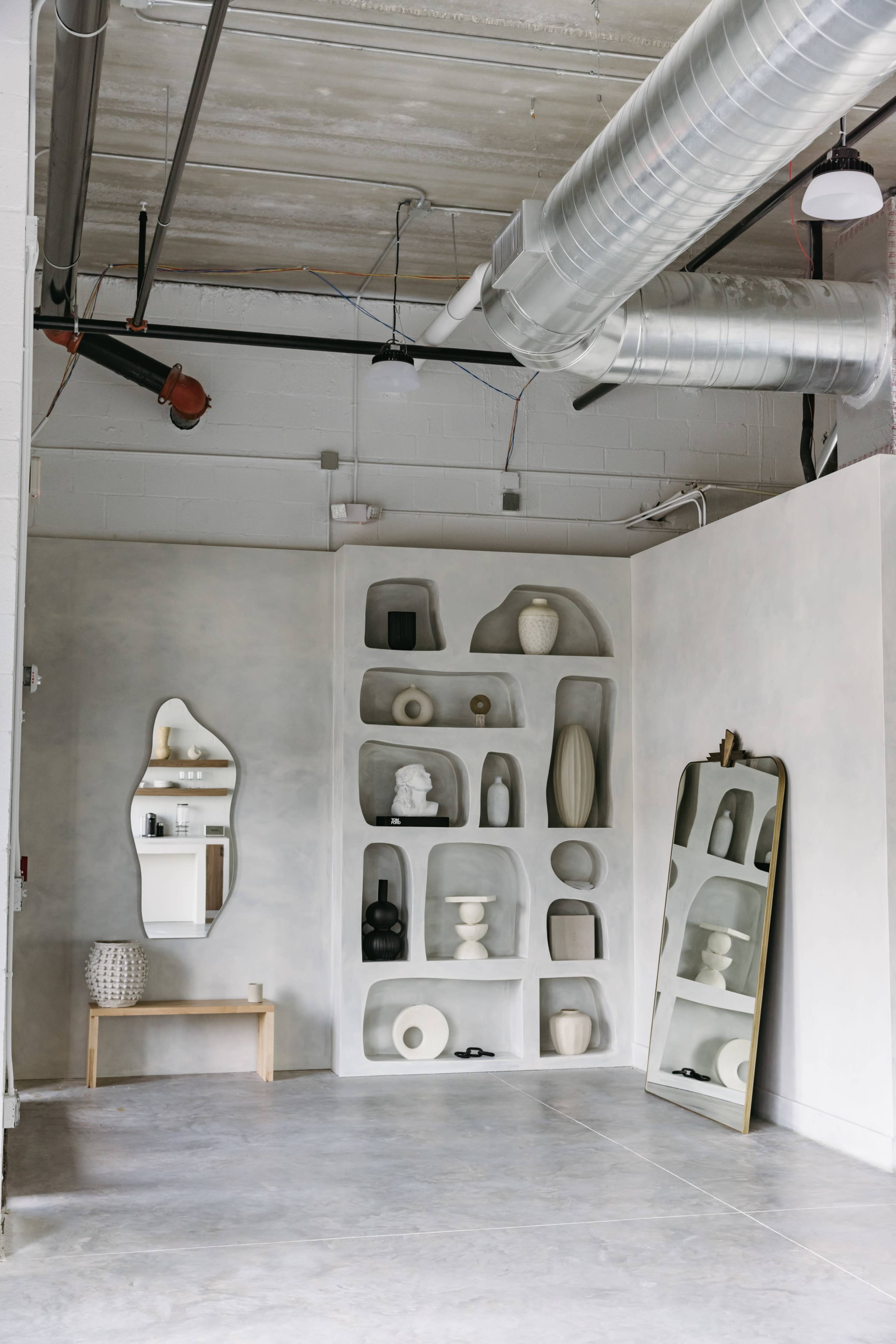 A minimalistic interior space featuring a textured gray wall with built-in shelves displaying various decorative vases and objects, alongside a tall mirror and a small wooden bench.