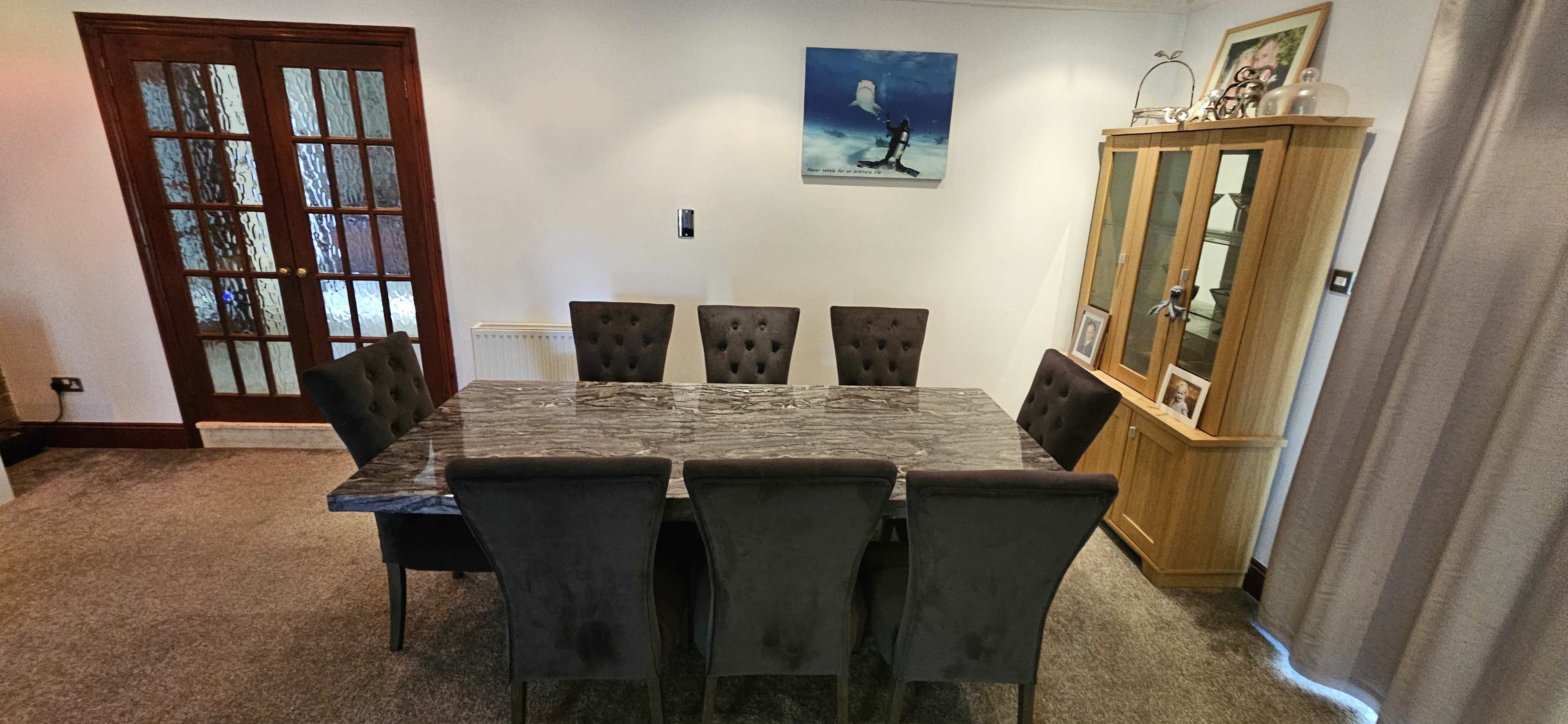 The image shows a dining room with a large marble table surrounded by six upholstered chairs, against a backdrop of a wooden cabinet and a painting on the wall.