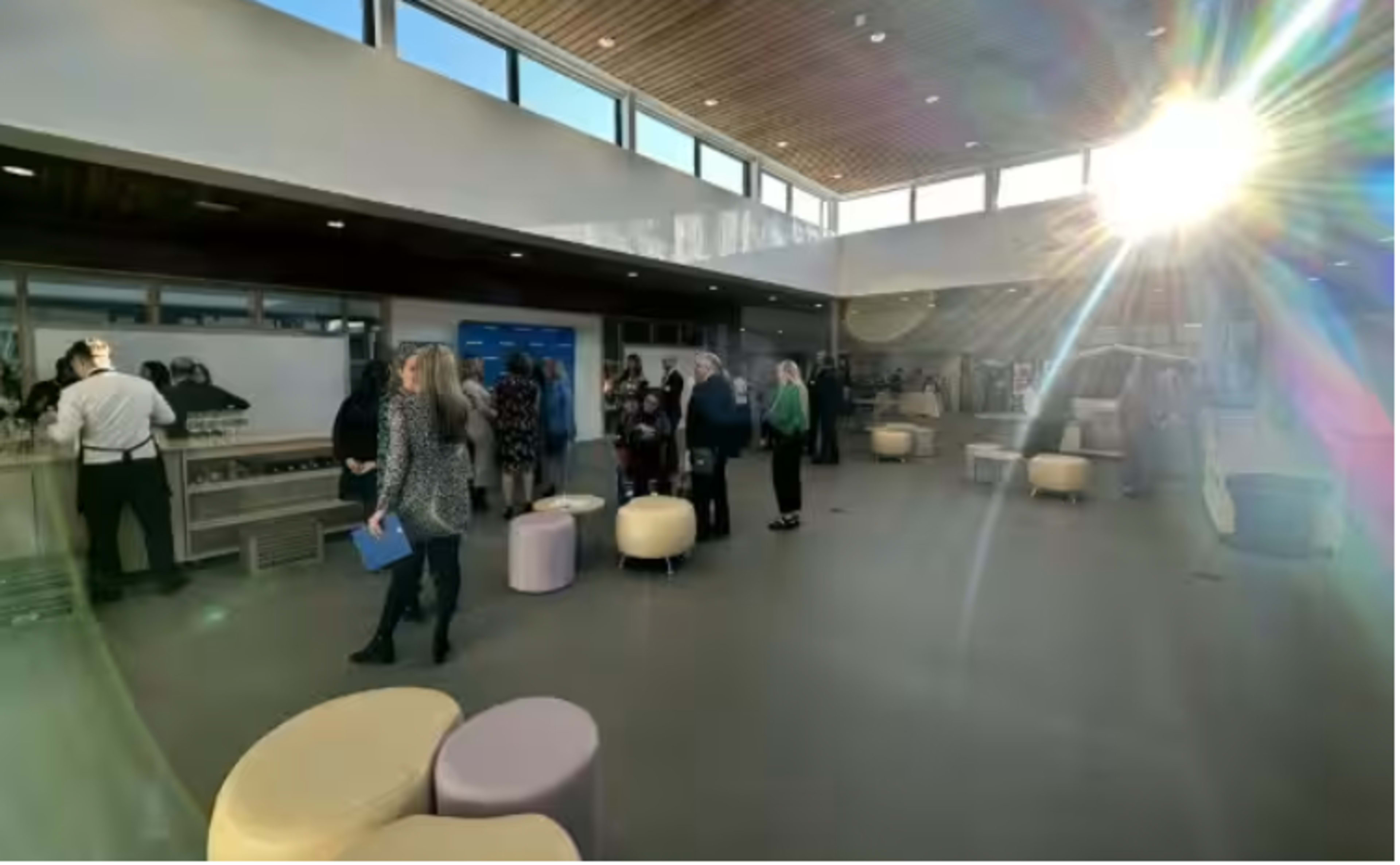 The image shows a crowded indoor space with people mingling, a service area on the left, and large windows allowing sunlight to stream in.