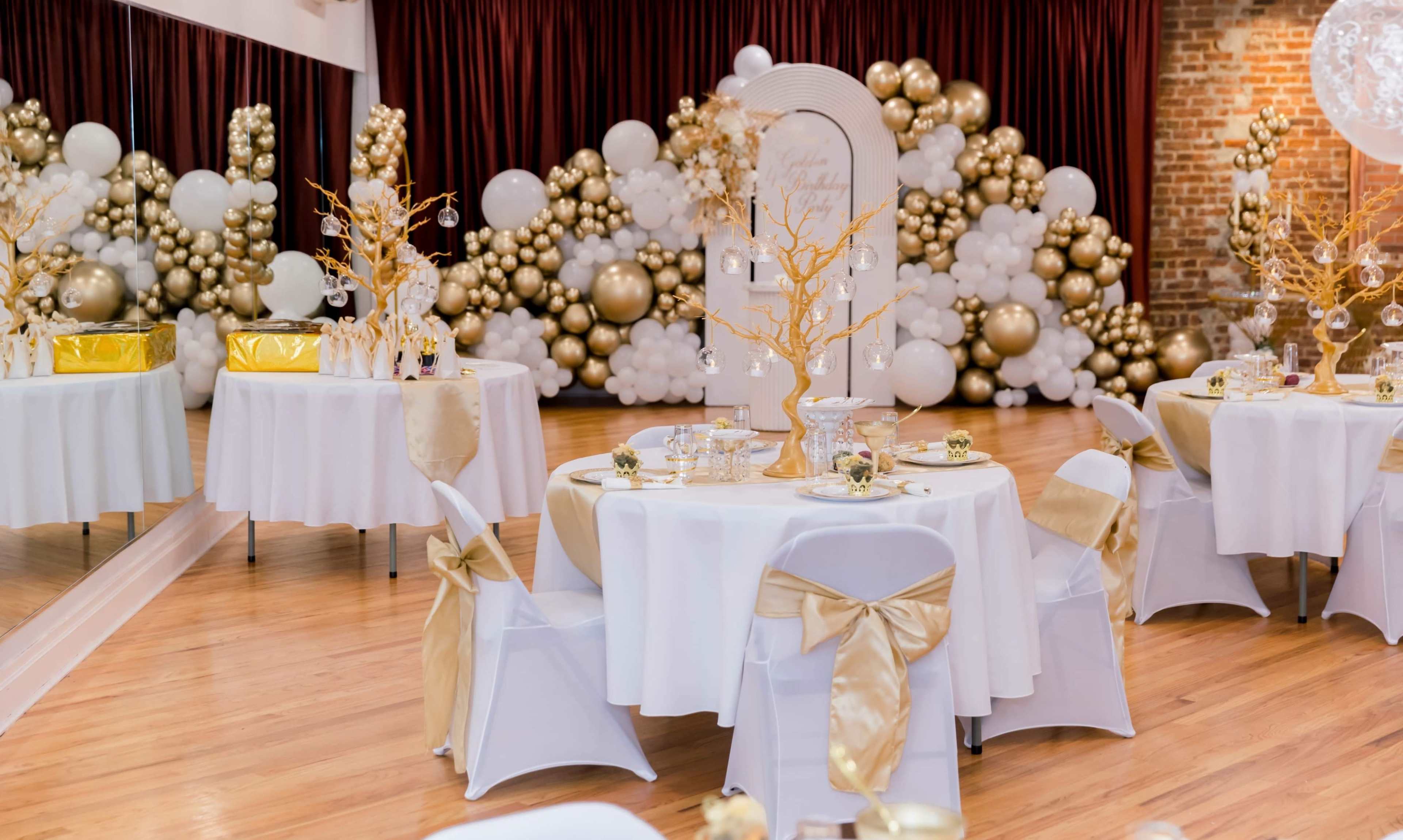 The image shows a decorated event space featuring round tables with white tablecloths and gold accents, surrounded by a backdrop of gold and white balloon arrangements.