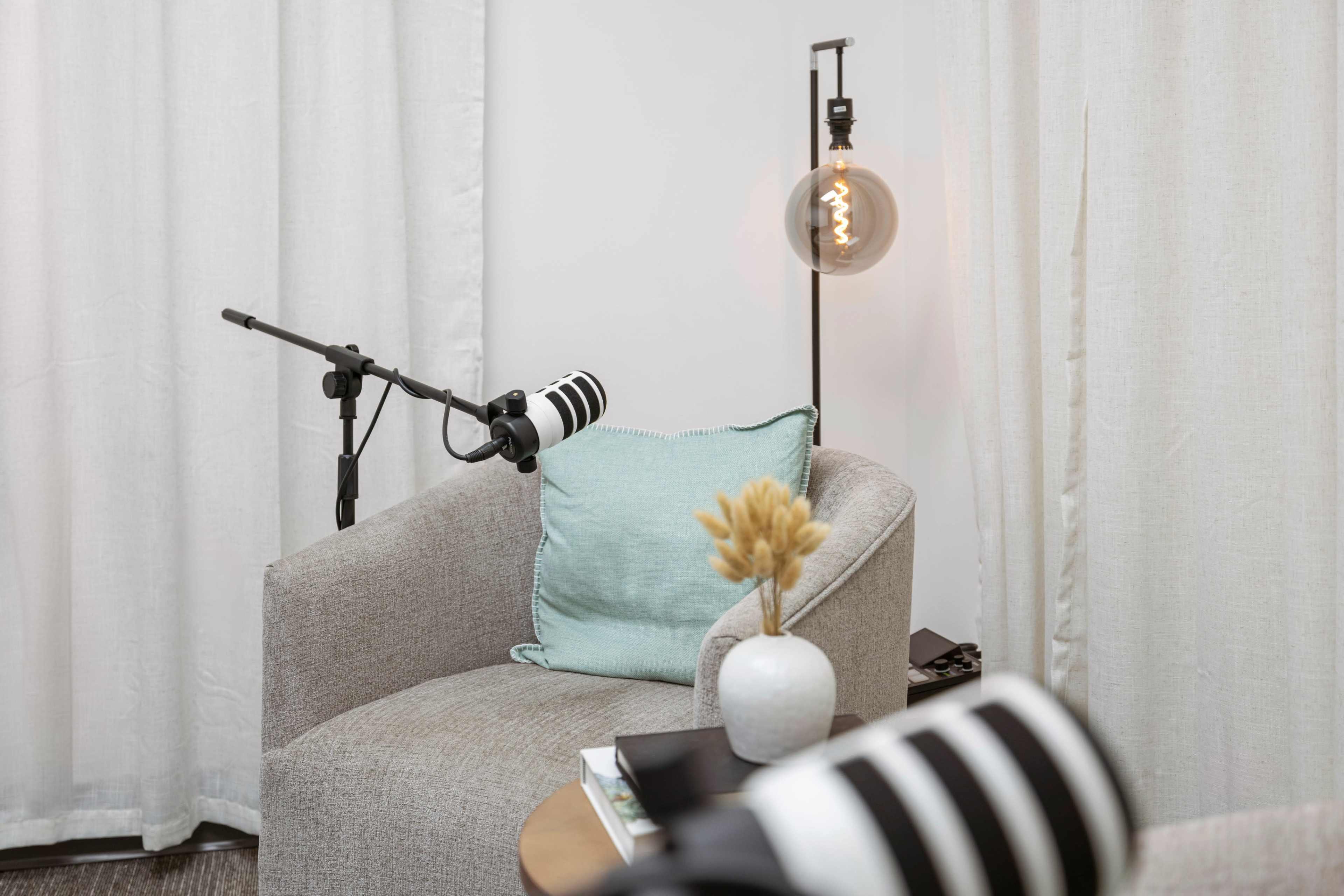 A microphone setup is positioned next to a cozy chair adorned with a light blue pillow, accompanied by a small vase of dried flowers and a lamp with an exposed bulb.