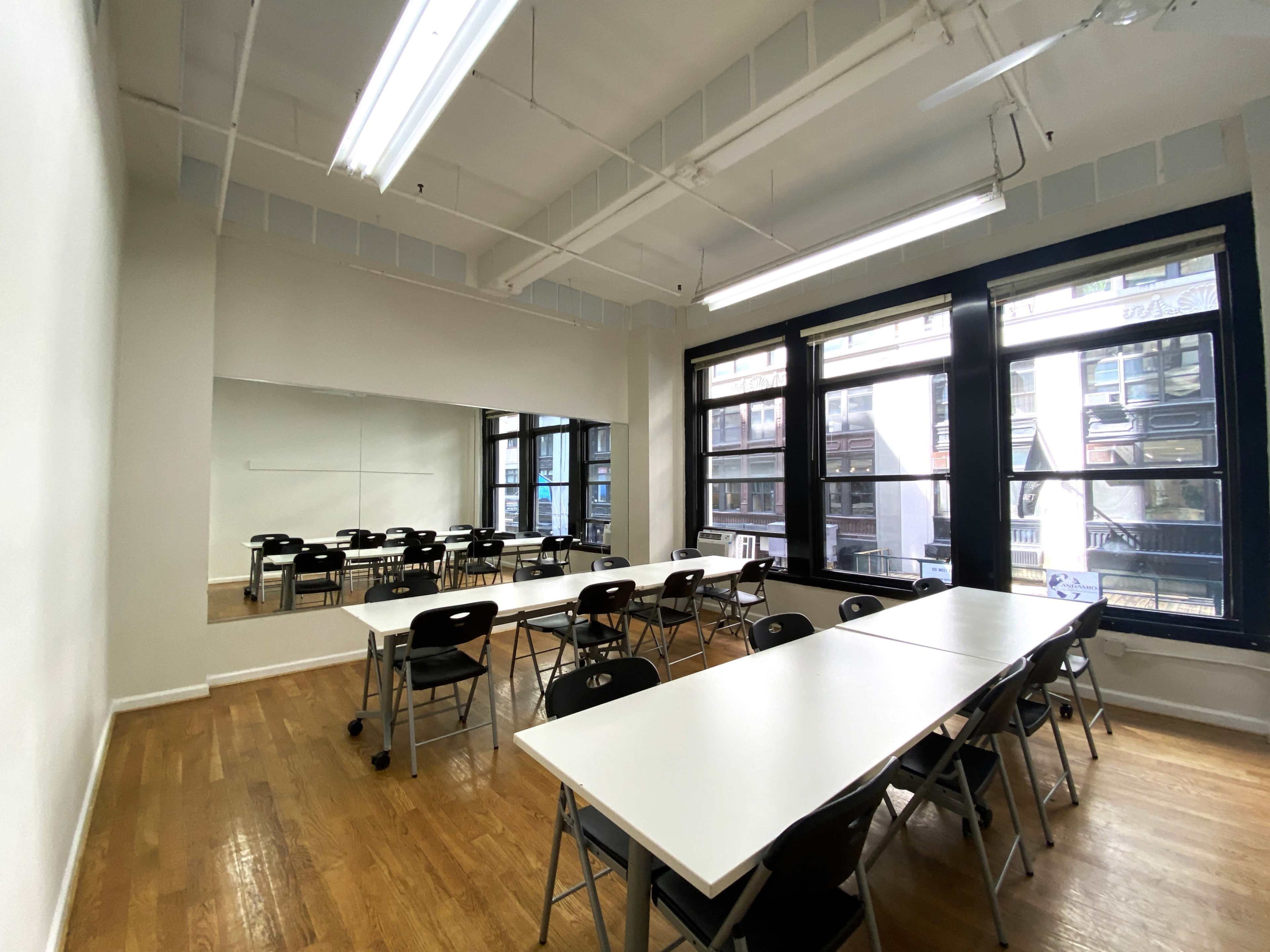 A spacious classroom with rows of white tables and black chairs, large windows providing natural light, and a wall mirror.