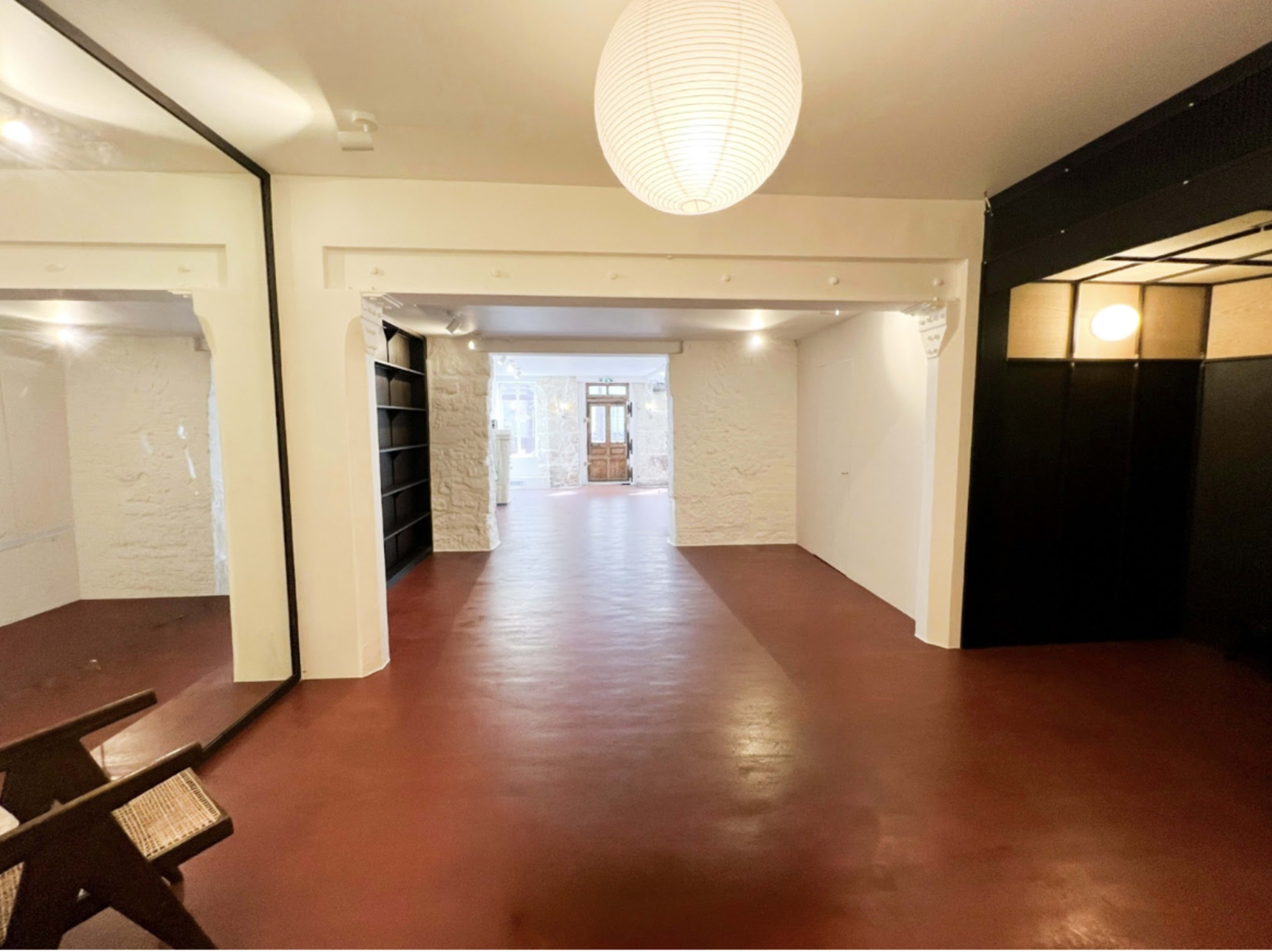The image shows a spacious, open room with a reddish floor, white walls, and a large mirror on one side, leading to a doorway framed by wooden doors.