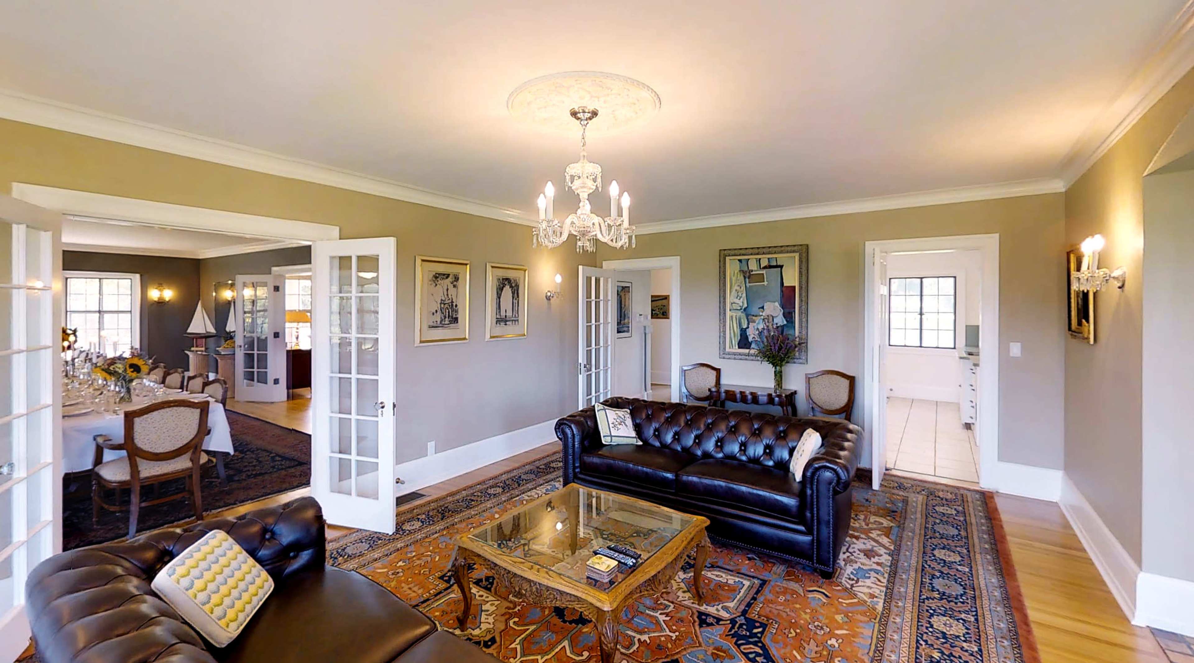 The living room features leather sofas, a glass coffee table, and a chandelier, with French doors leading to an adjoining dining area.