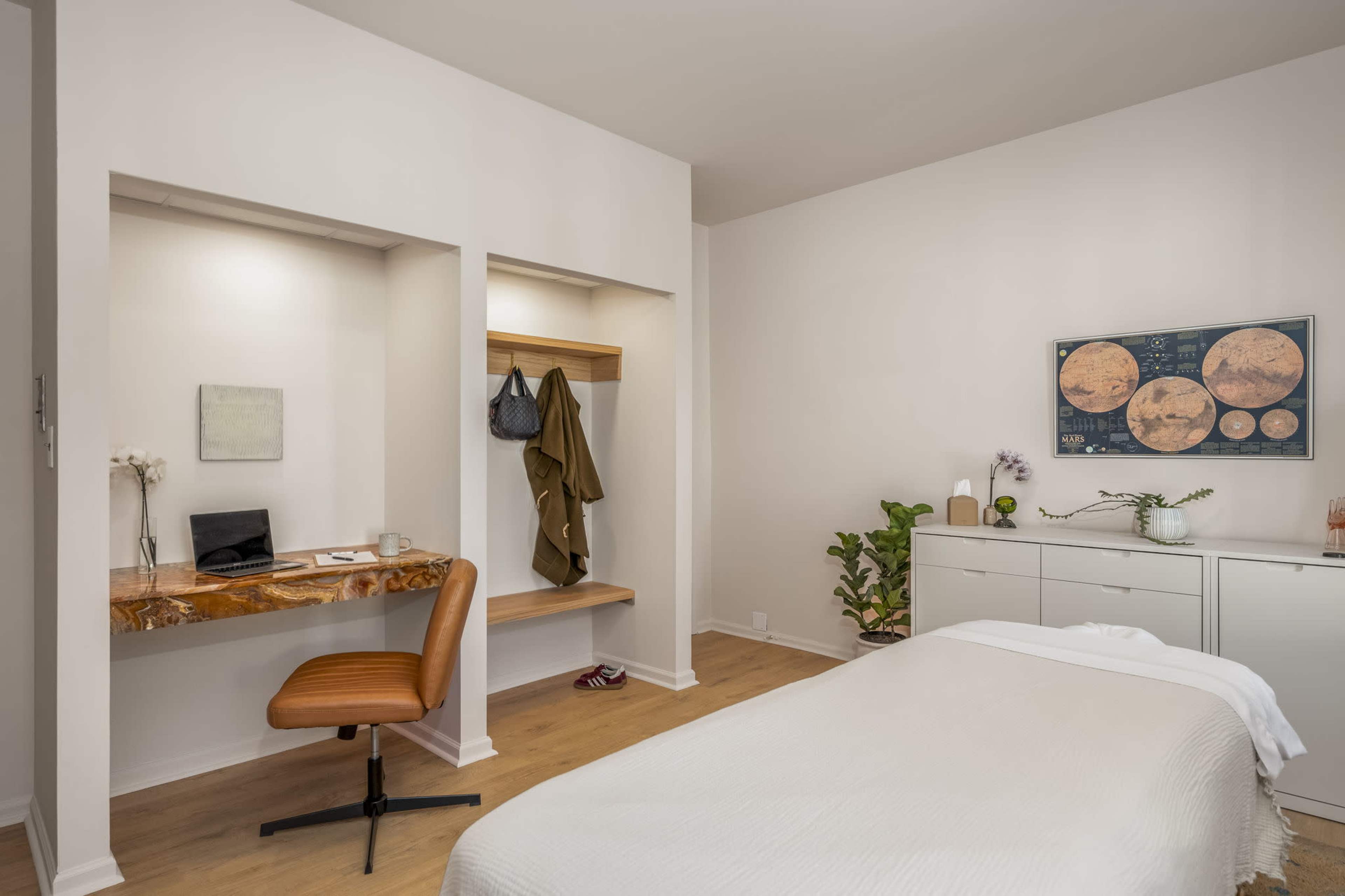 The image shows a minimalist bedroom with a bed, a small desk area with a chair, and a rack for hanging clothes, along with a potted plant.
