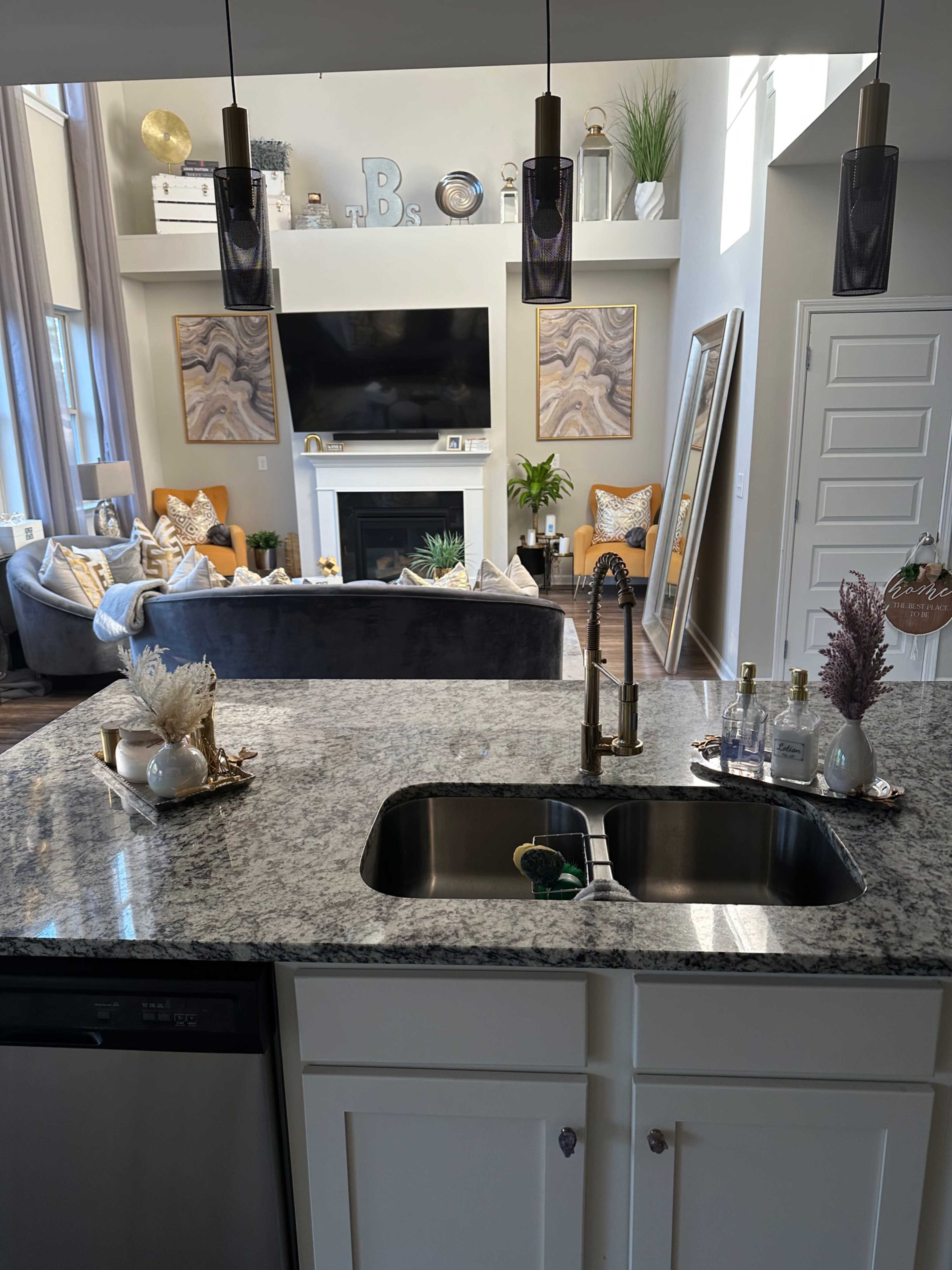 A modern kitchen with a granite countertop and double sink, overlooking a spacious living room that features a large television, decorative plants, and comfortable seating.