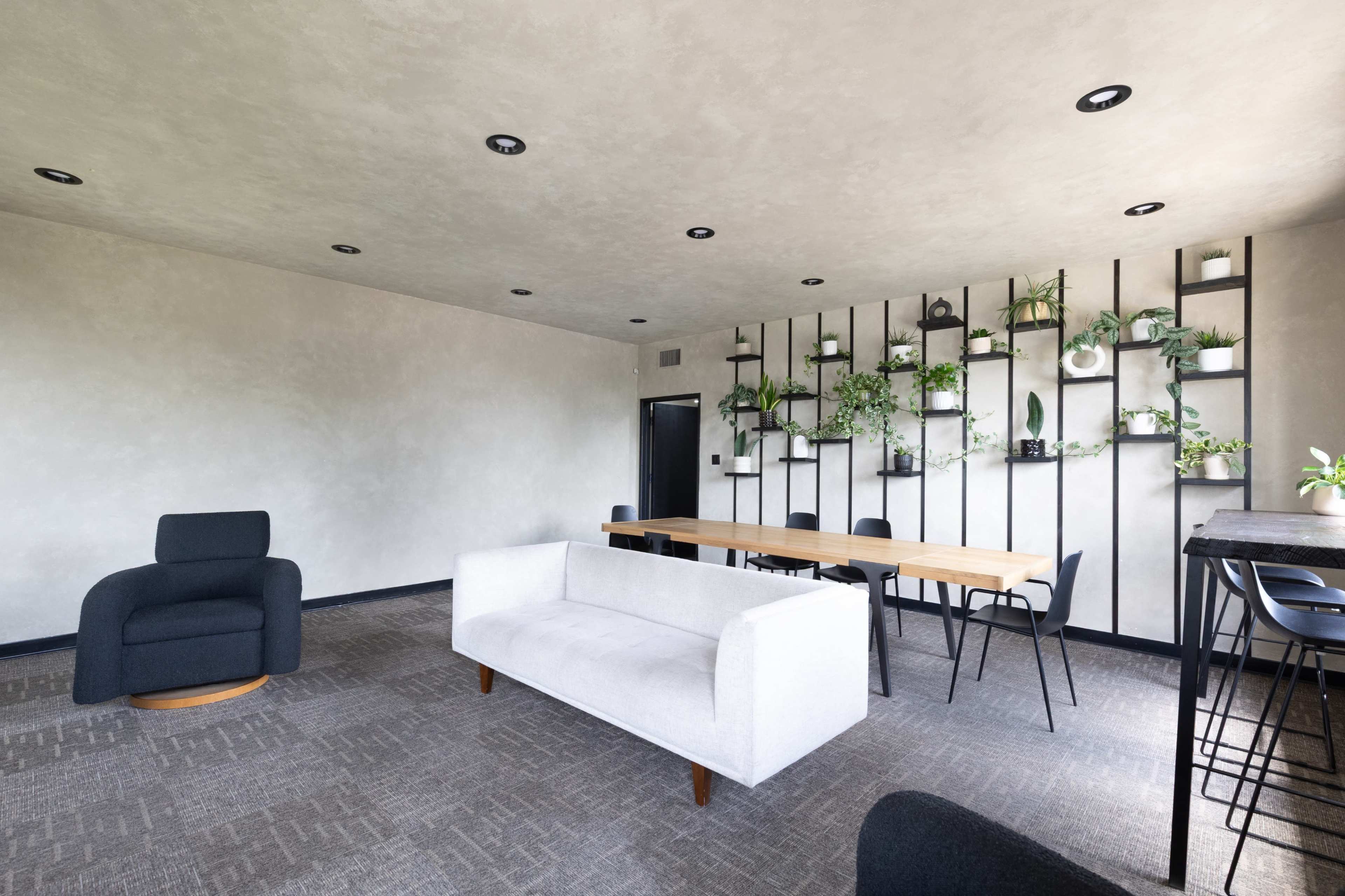 The image shows a modern, minimalist meeting room with a white sofa, a wooden table surrounded by black chairs, and a plant-filled wall installation.