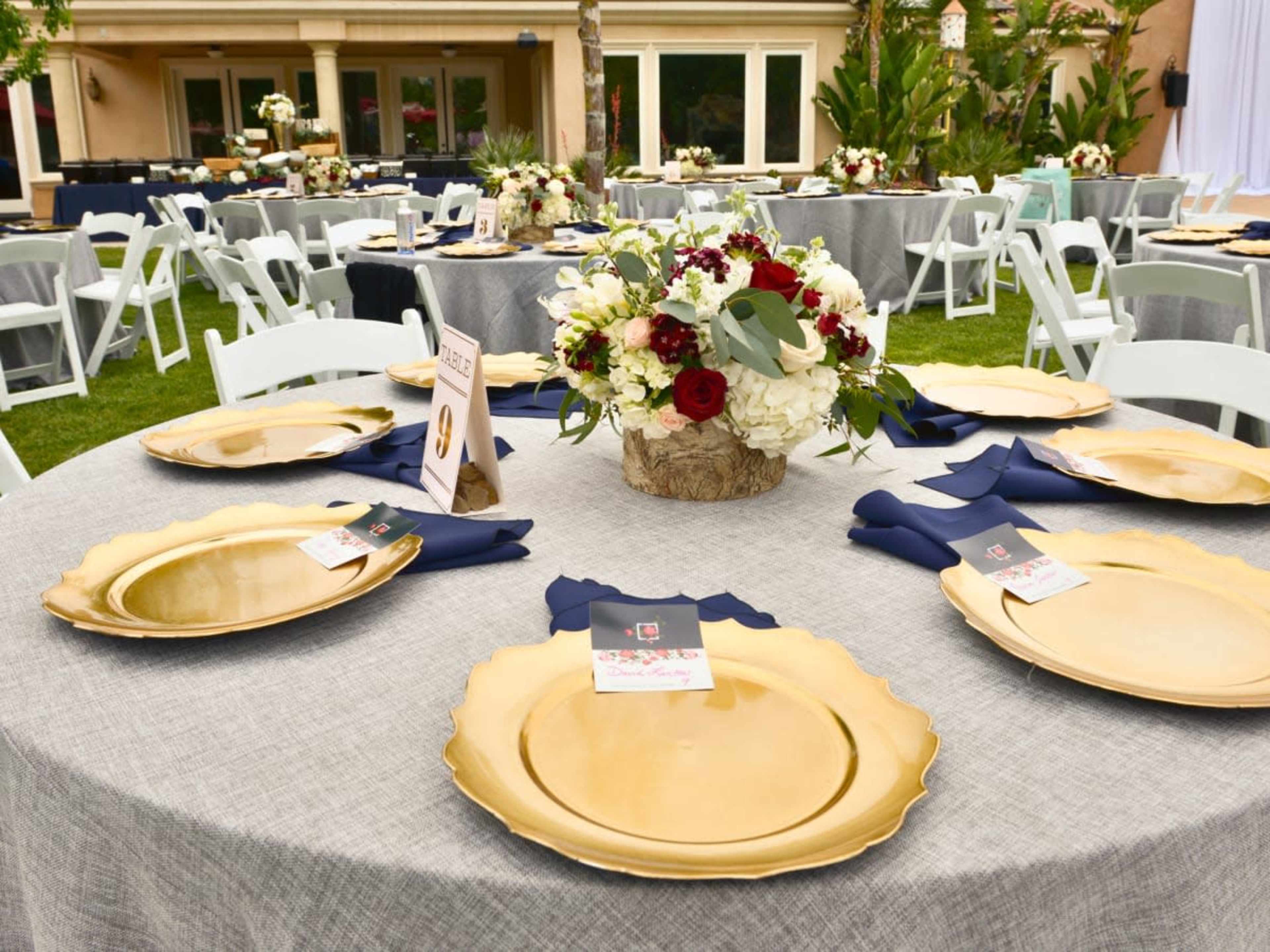The image shows a tablescape set outdoors featuring gold charger plates, floral centerpieces, and navy blue napkins arranged atop gray tablecloths.