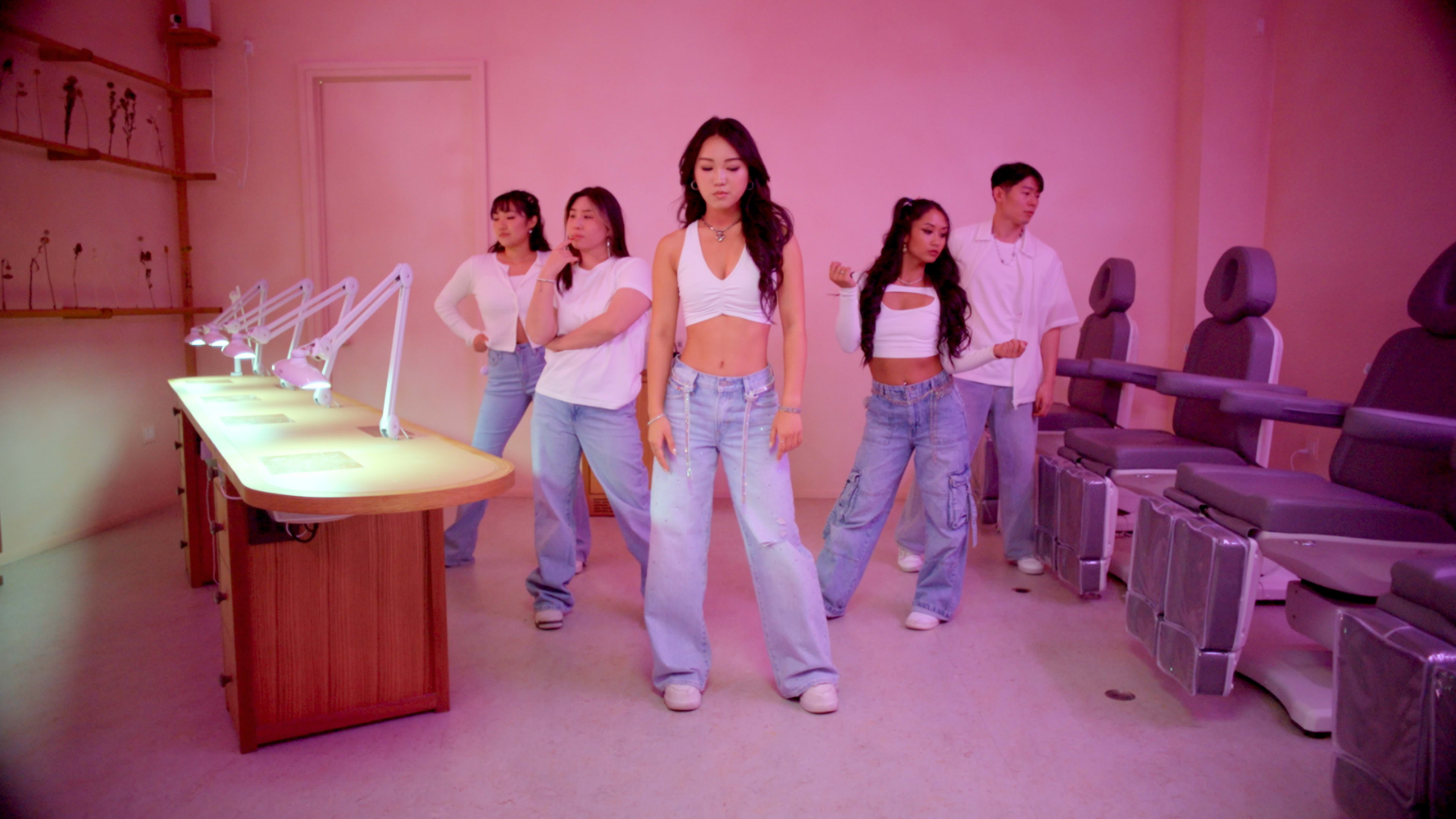 A group of five individuals dressed in casual clothing is performing a coordinated dance in a brightly lit room with pink walls.