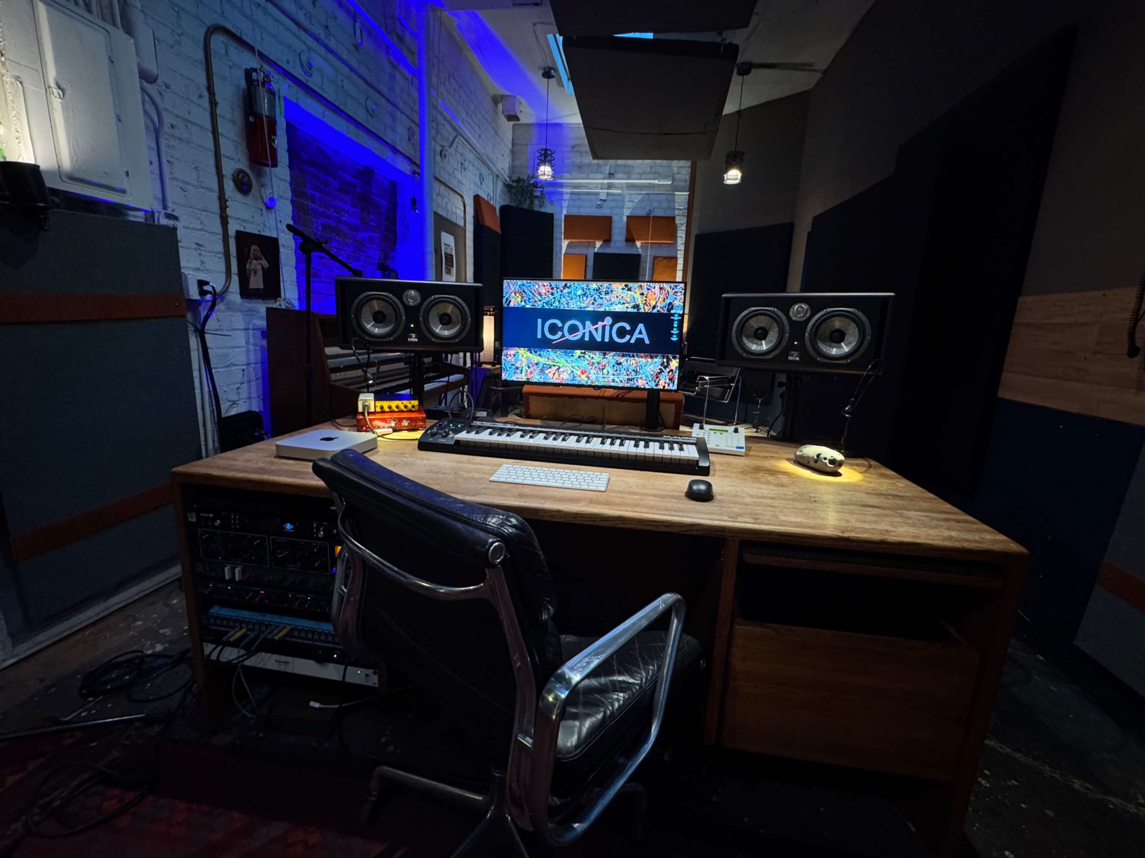 The image shows a modern music production studio featuring a wooden desk with electronic instruments, computer monitors, and sound equipment, illuminated by ambient lighting.