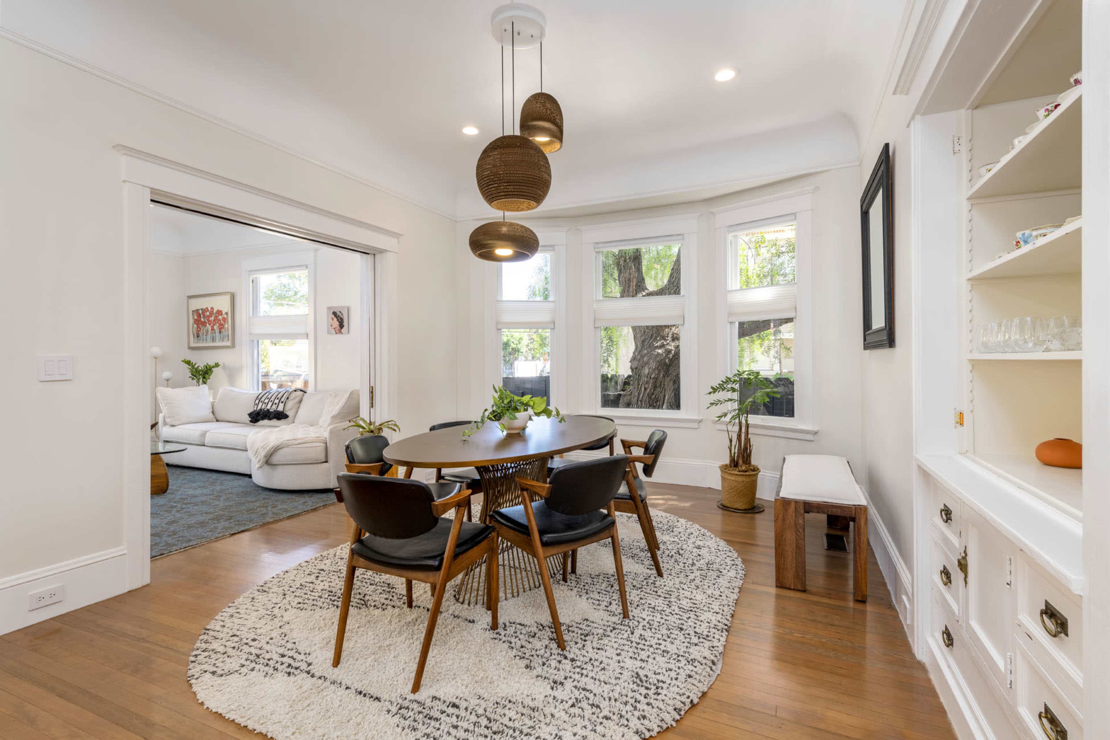A dining area features a round table surrounded by dark chairs, positioned on a circular rug, with large windows allowing natural light and a view of a cozy living room.