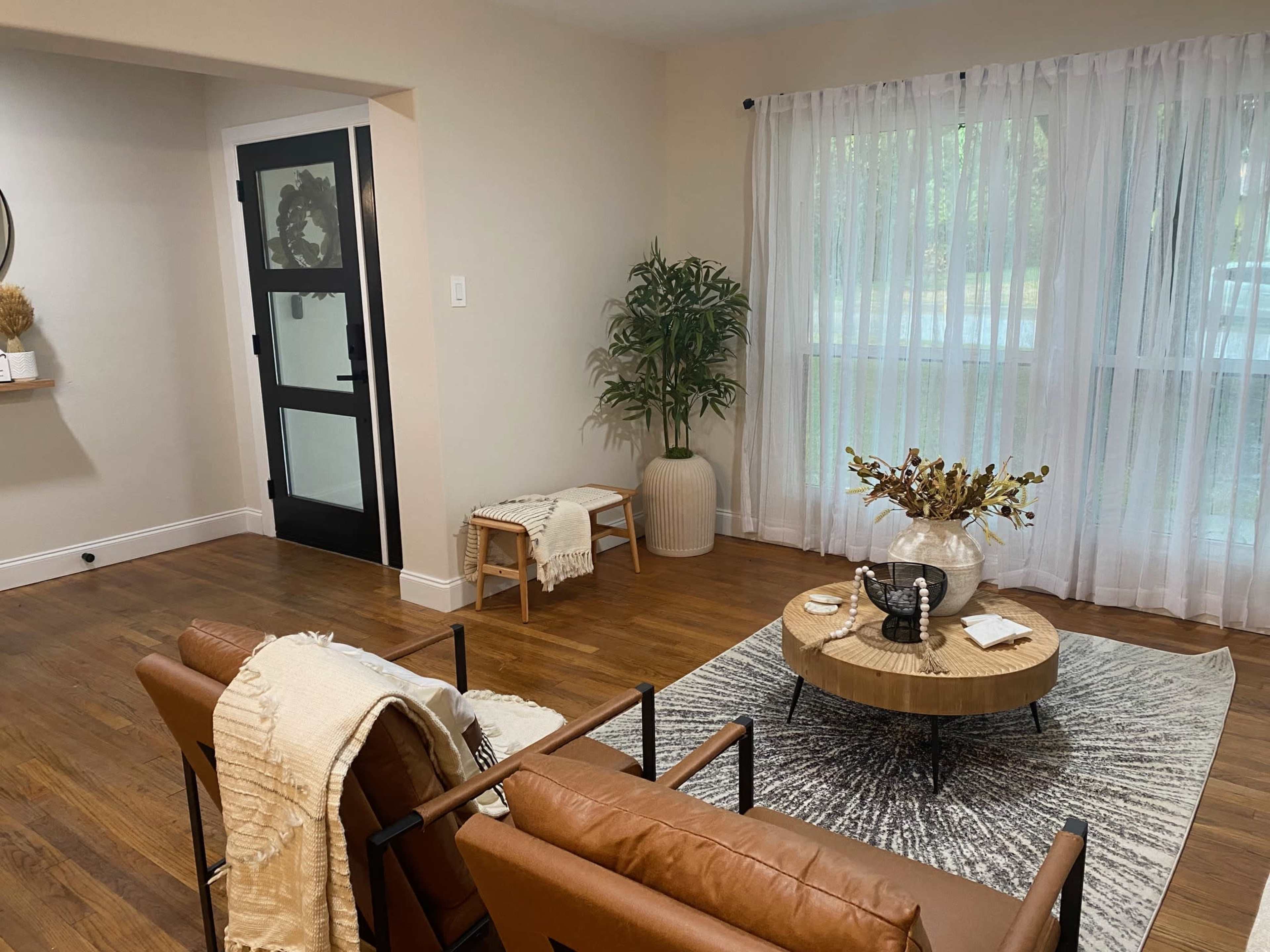 A cozy living room features a brown leather sofa, a round wooden coffee table on a patterned rug, and large windows with sheer curtains.